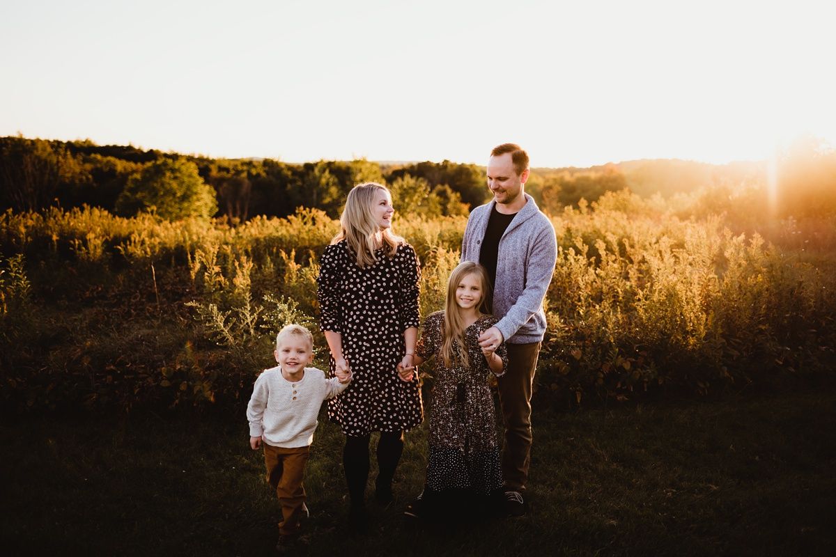 Family of four poses in golden field during sunset wearing polka dot dress and casual attire.