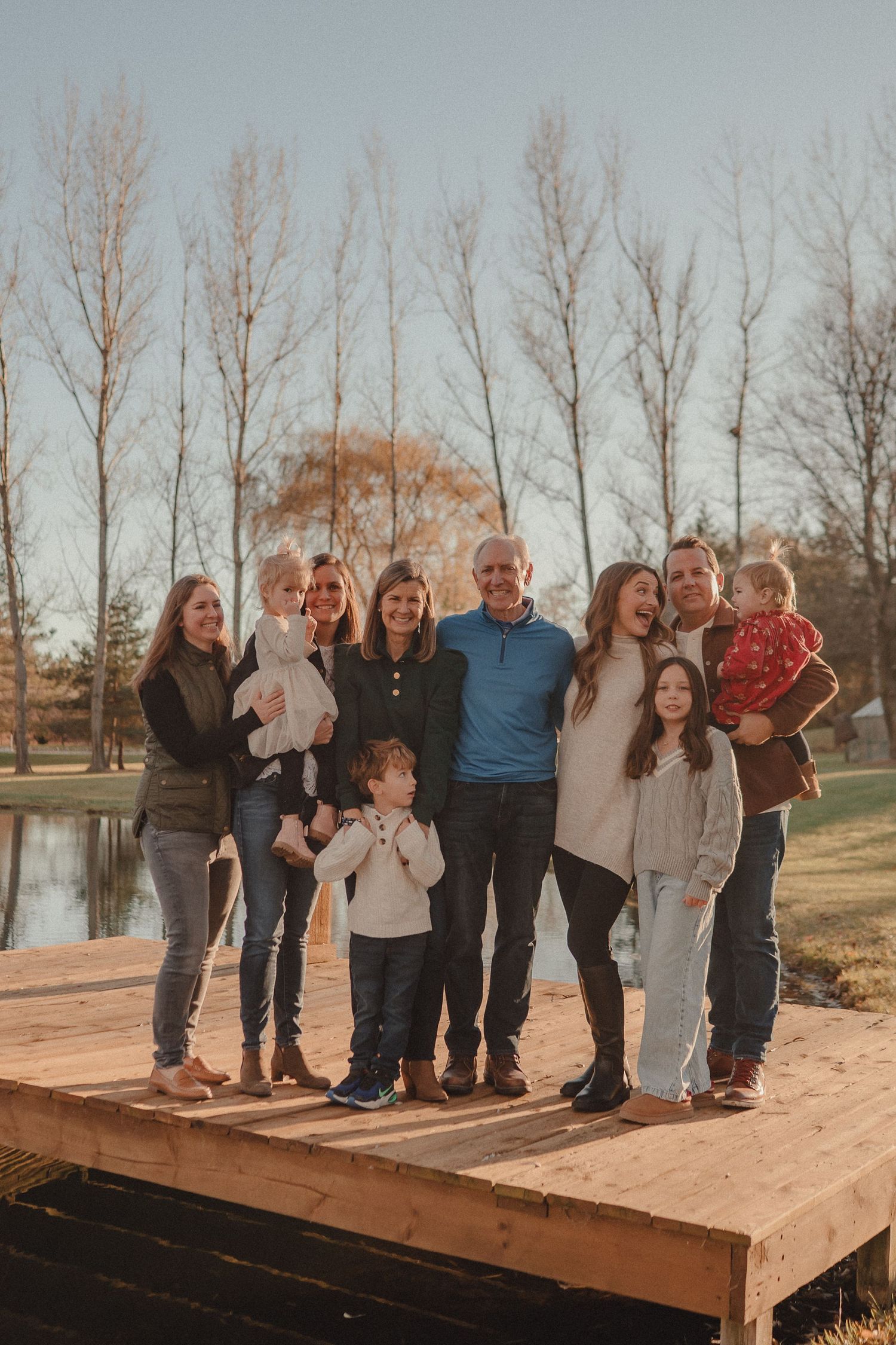 A large group poses together on a wooden dock by a lake during autumn with bare trees in the background.