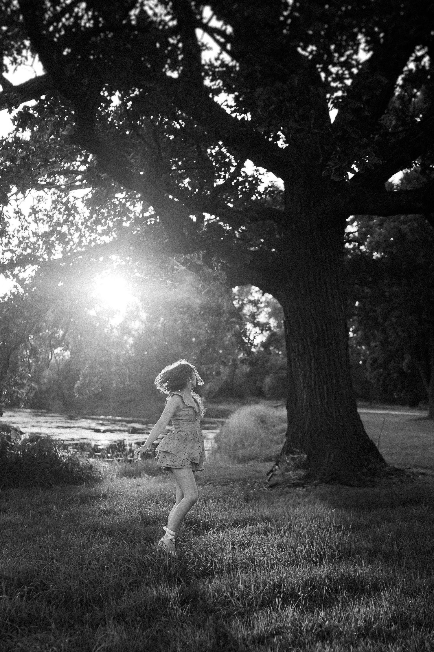 Black and white photo of a person dancing joyfully under a large tree with sunlight streaming through branches.