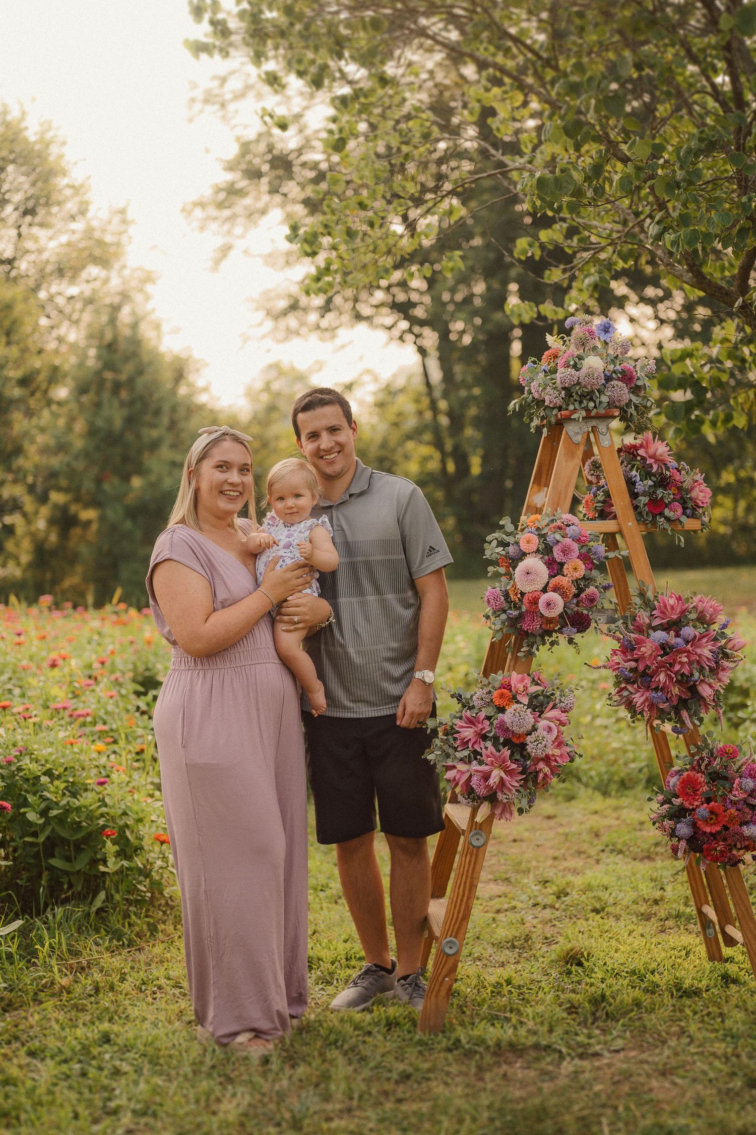Family posing next to a wooden triangular flower stand decorated with pink blooms in an outdoor garden setting.