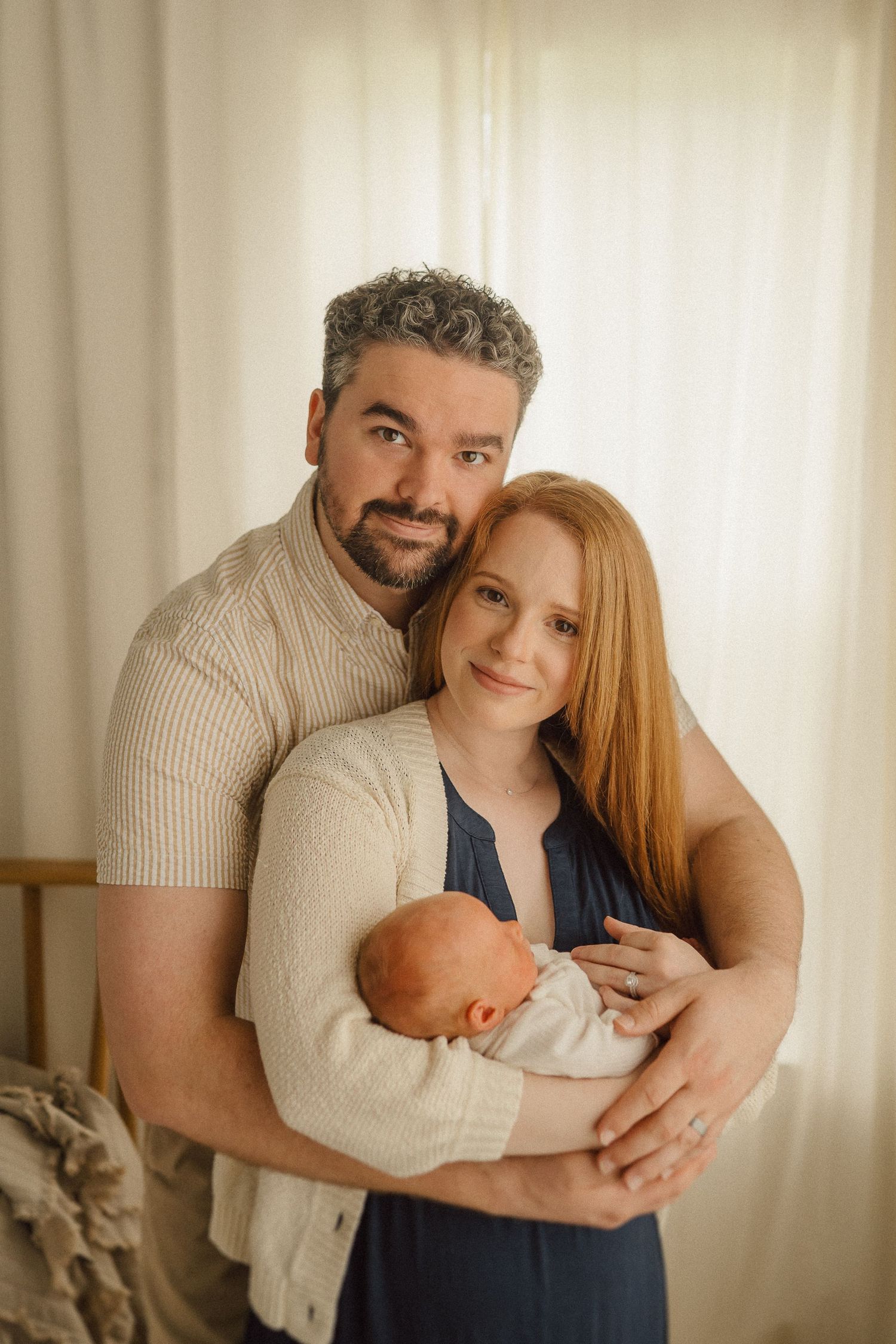 A loving family portrait showing parents holding their newborn baby while standing by a window with soft lighting.
