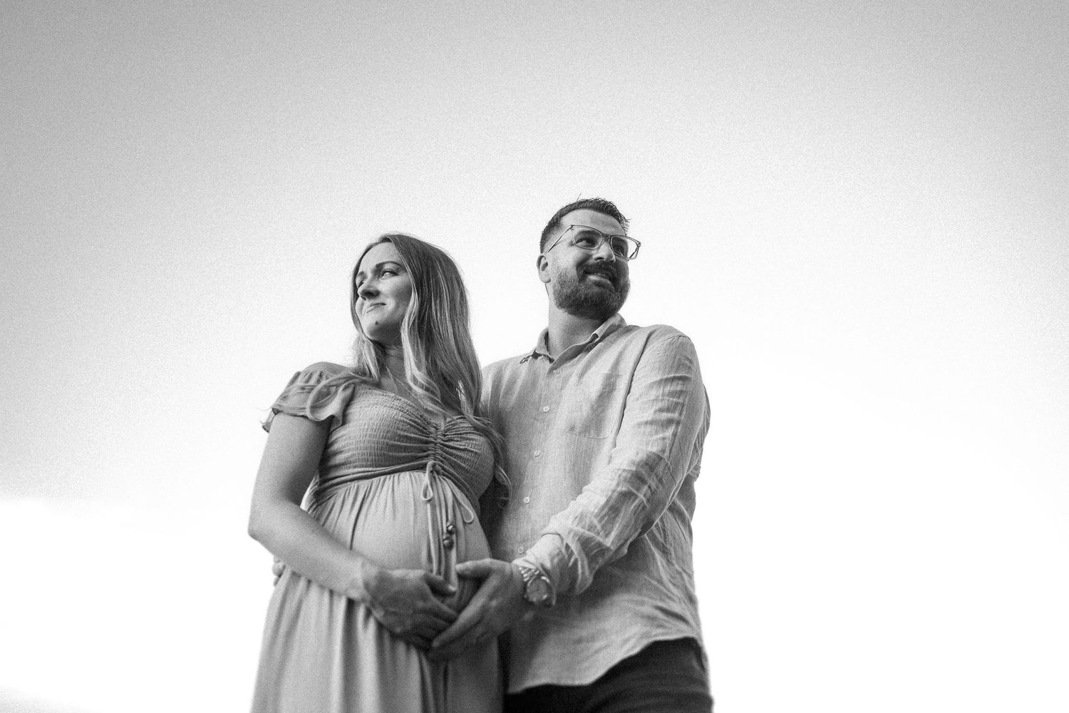 Black and white low angle portrait of a couple posing together outdoors, with the woman in a dress and man in light shirt.