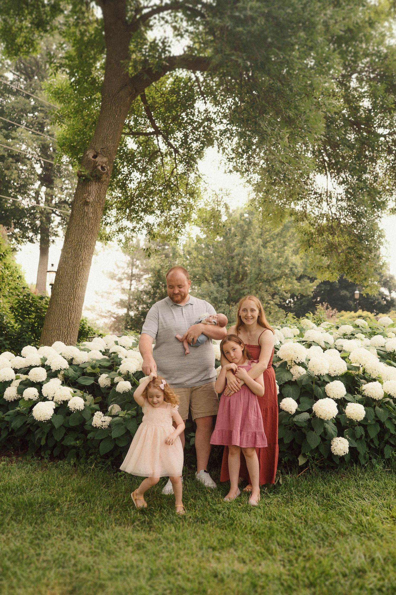 Family relaxes together on grassy hill with beautiful white hydrangea bushes in background.