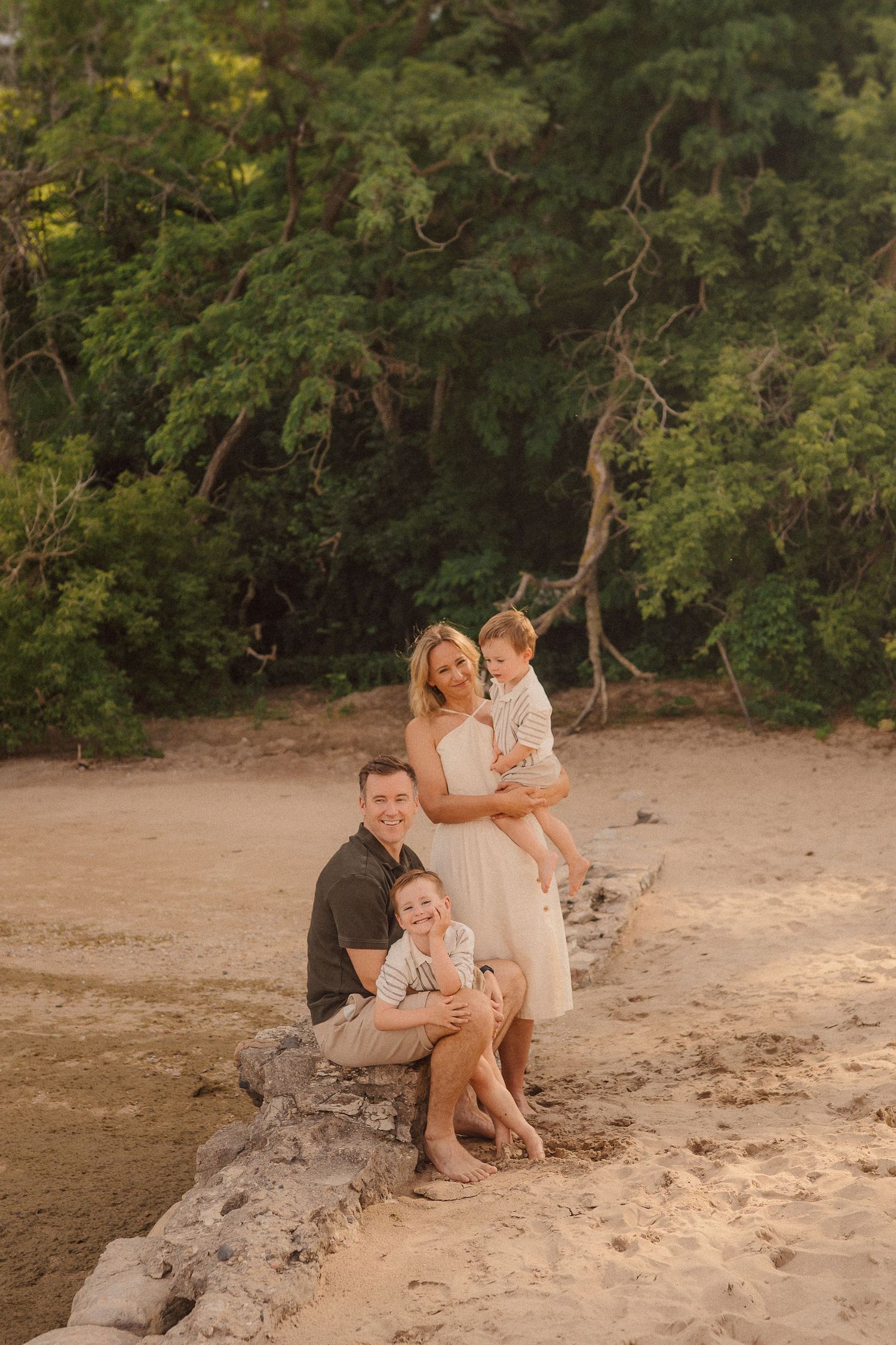 Family enjoys sunset at the beach while sitting together on driftwood near forest trees.