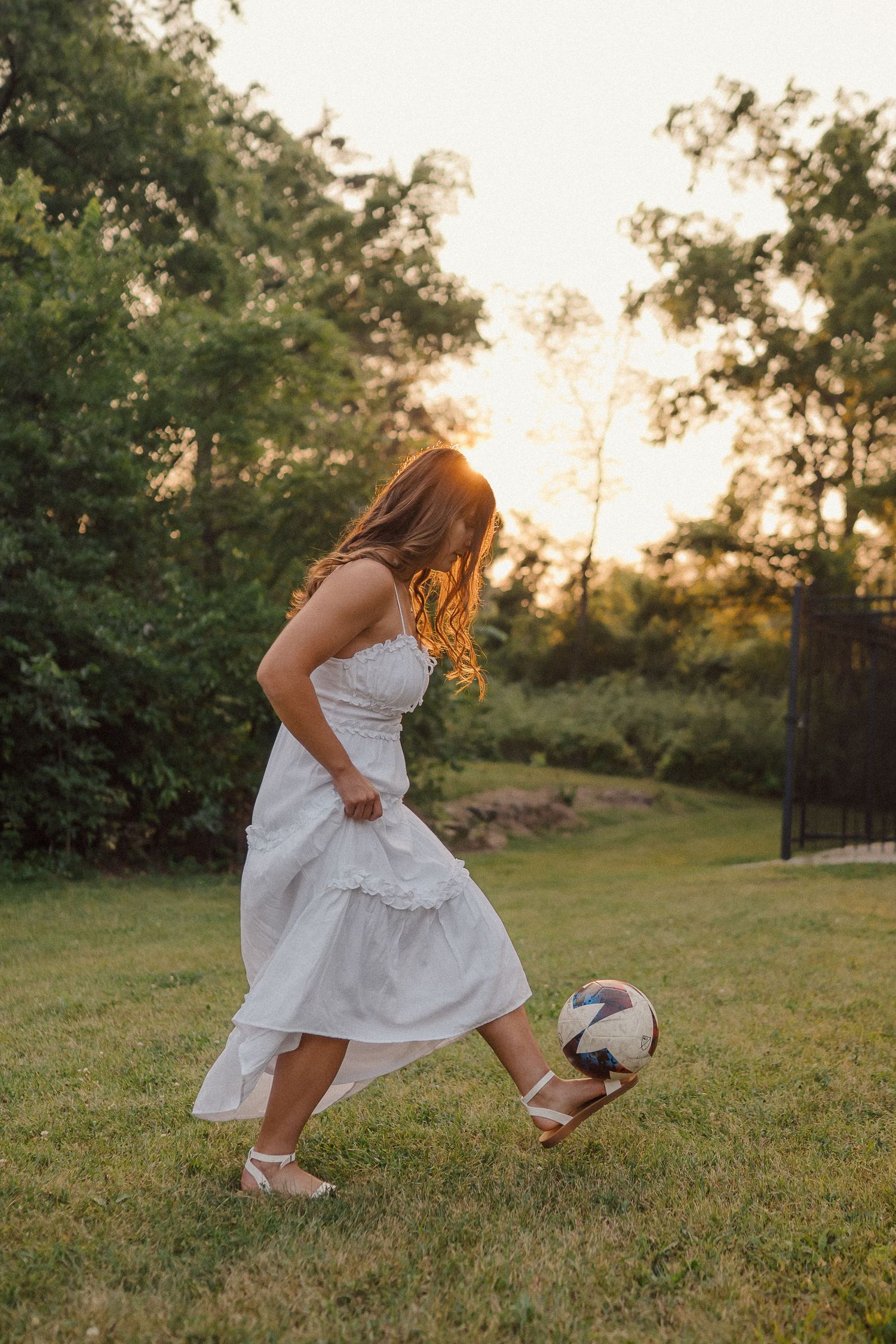 A person in a white dress kicks a soccer ball in a sunny garden at sunset.