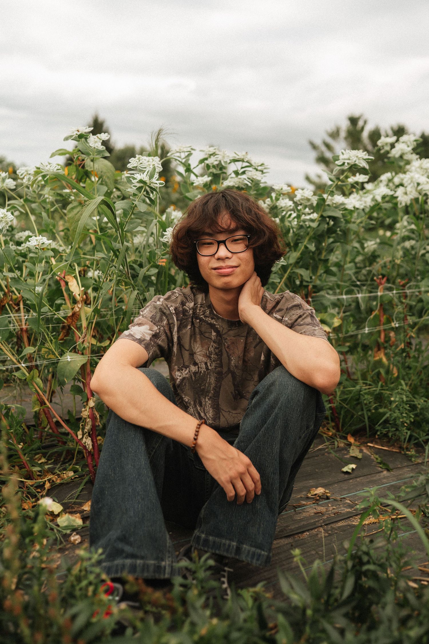 Someone in dark clothing and glasses poses casually in a garden with green plants and vines.
