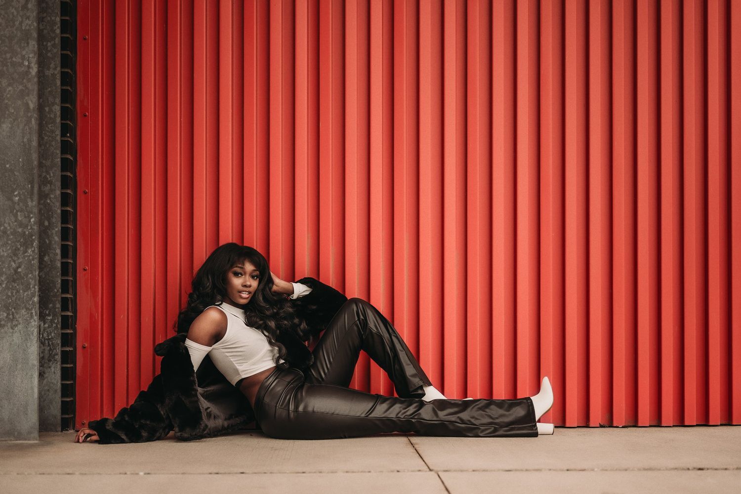 A model in leather pants and fur coat reclines against a red industrial backdrop.