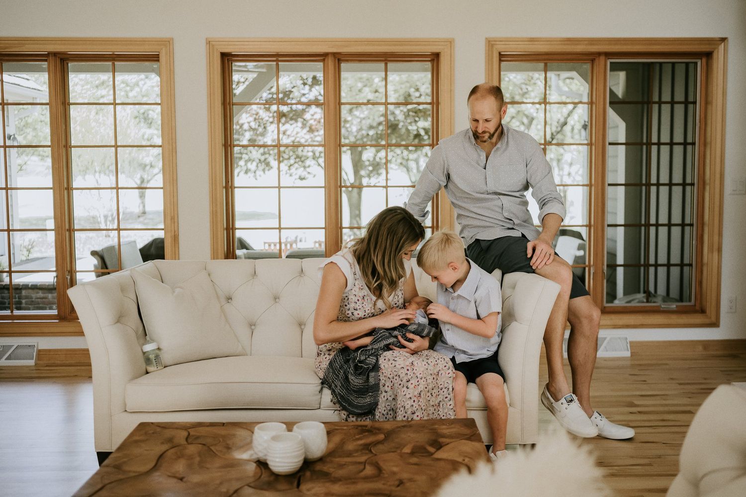 A family gathers on a cream-colored couch in front of large windows in a sunlit living room.