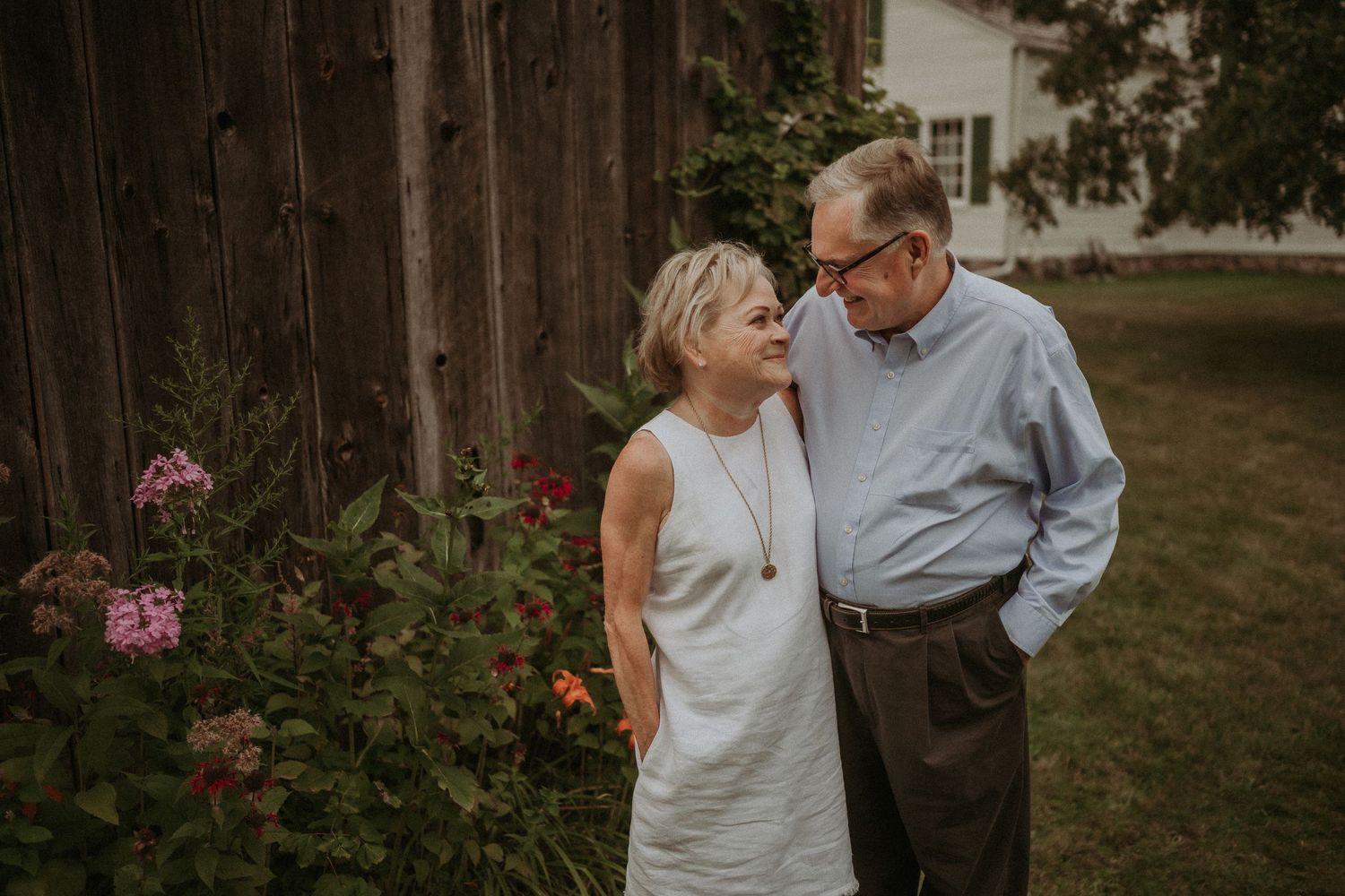 Outdoor portrait of a couple in light colored clothing standing by blooming garden flowers.