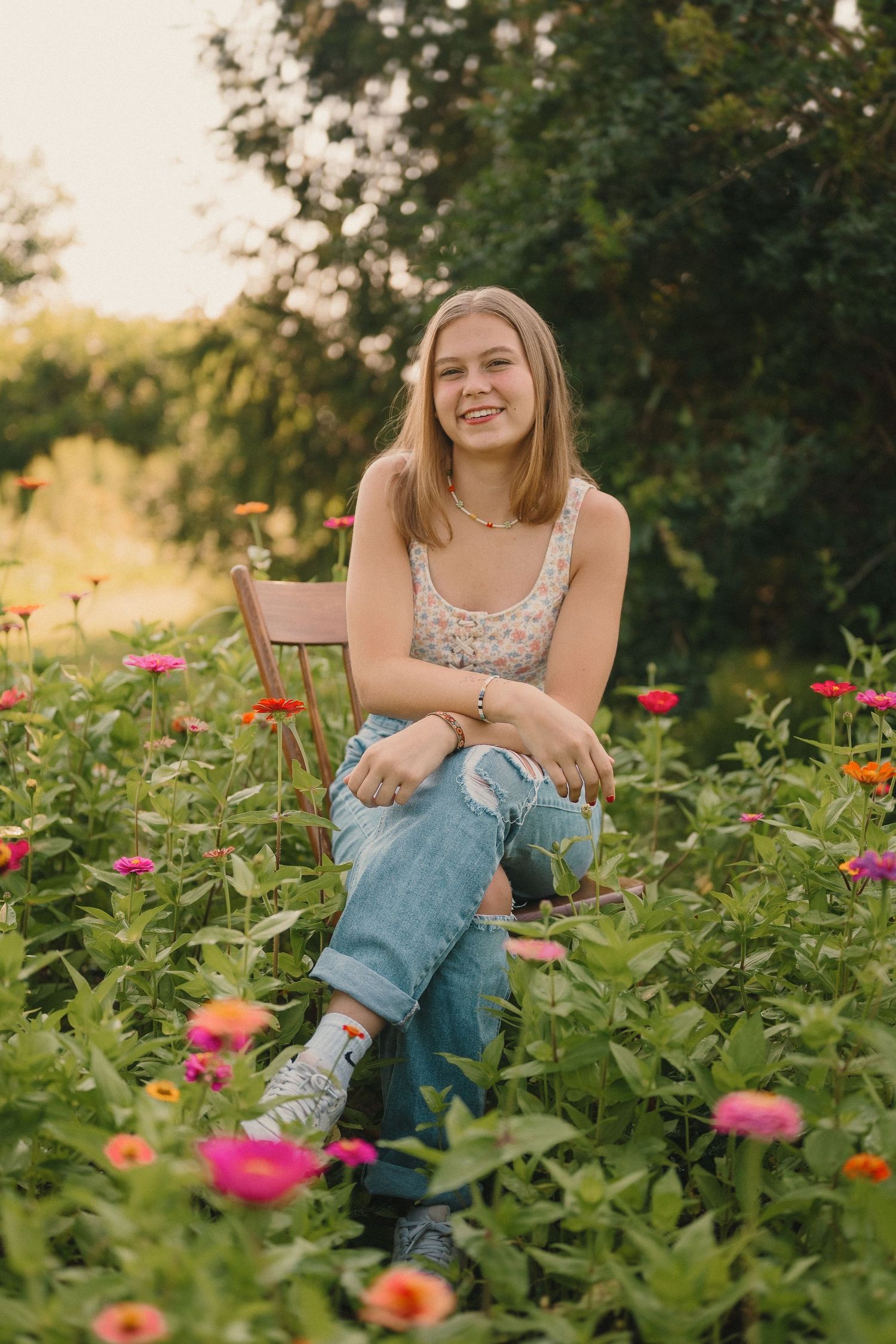 Person in a floral top and jeans sits among colorful flowers in a garden at sunset.
