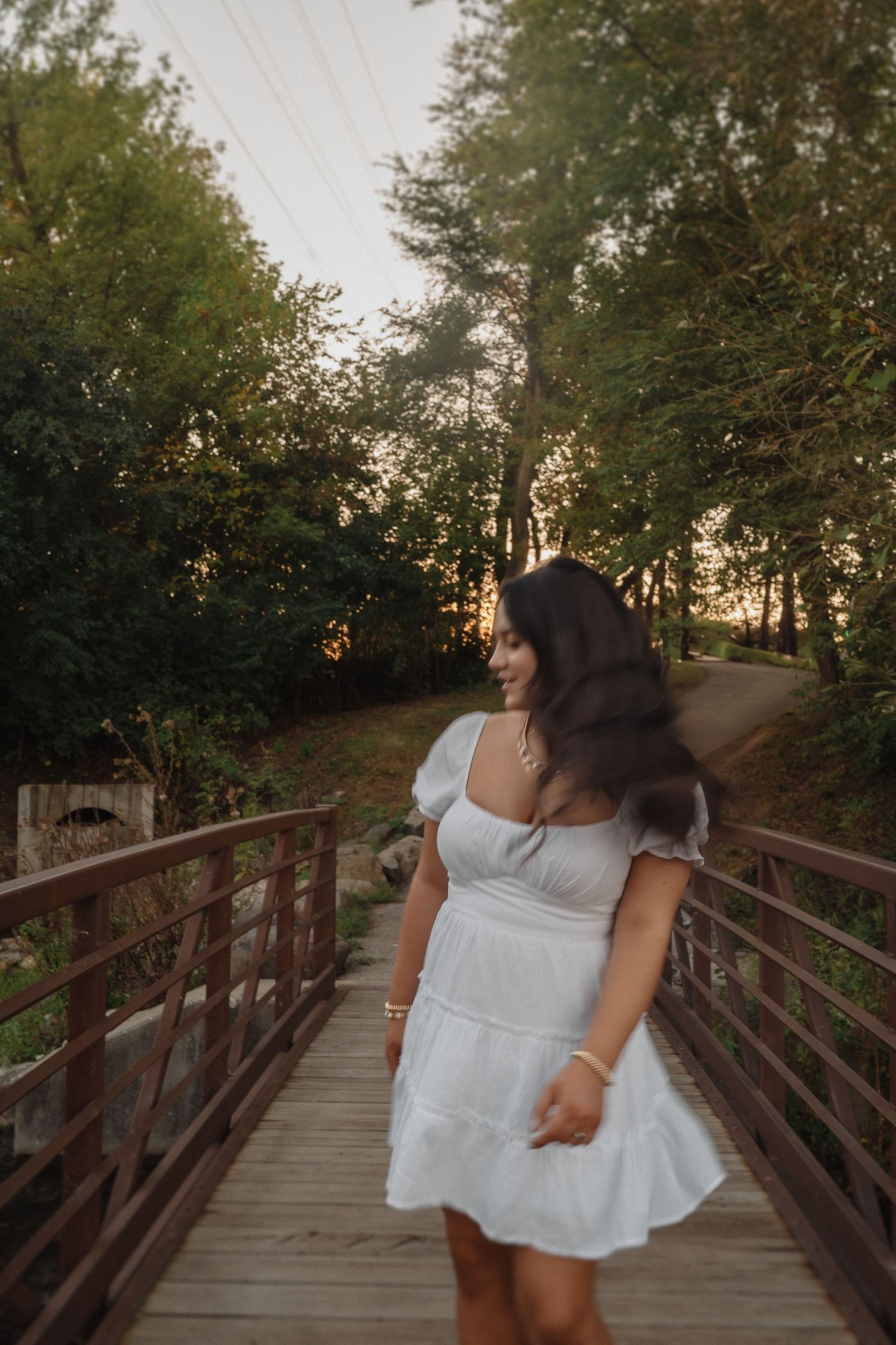 A person in a white dress stands on a rustic wooden bridge pathway amid lush greenery at dusk.