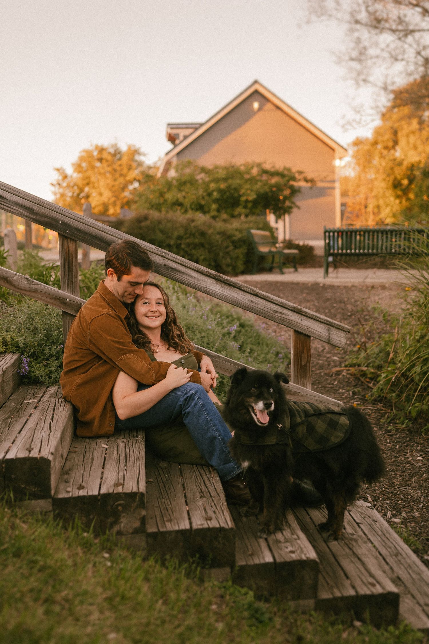A couple embraces on wooden steps with their black dog during a warm autumn afternoon.
