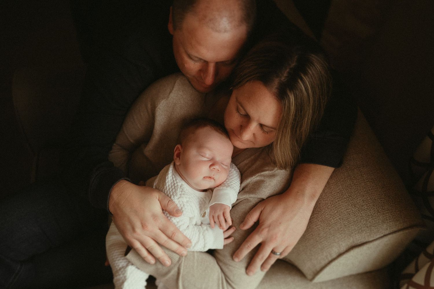 A tender family moment in moody lighting shows parents embracing their newborn baby in an intimate setting.