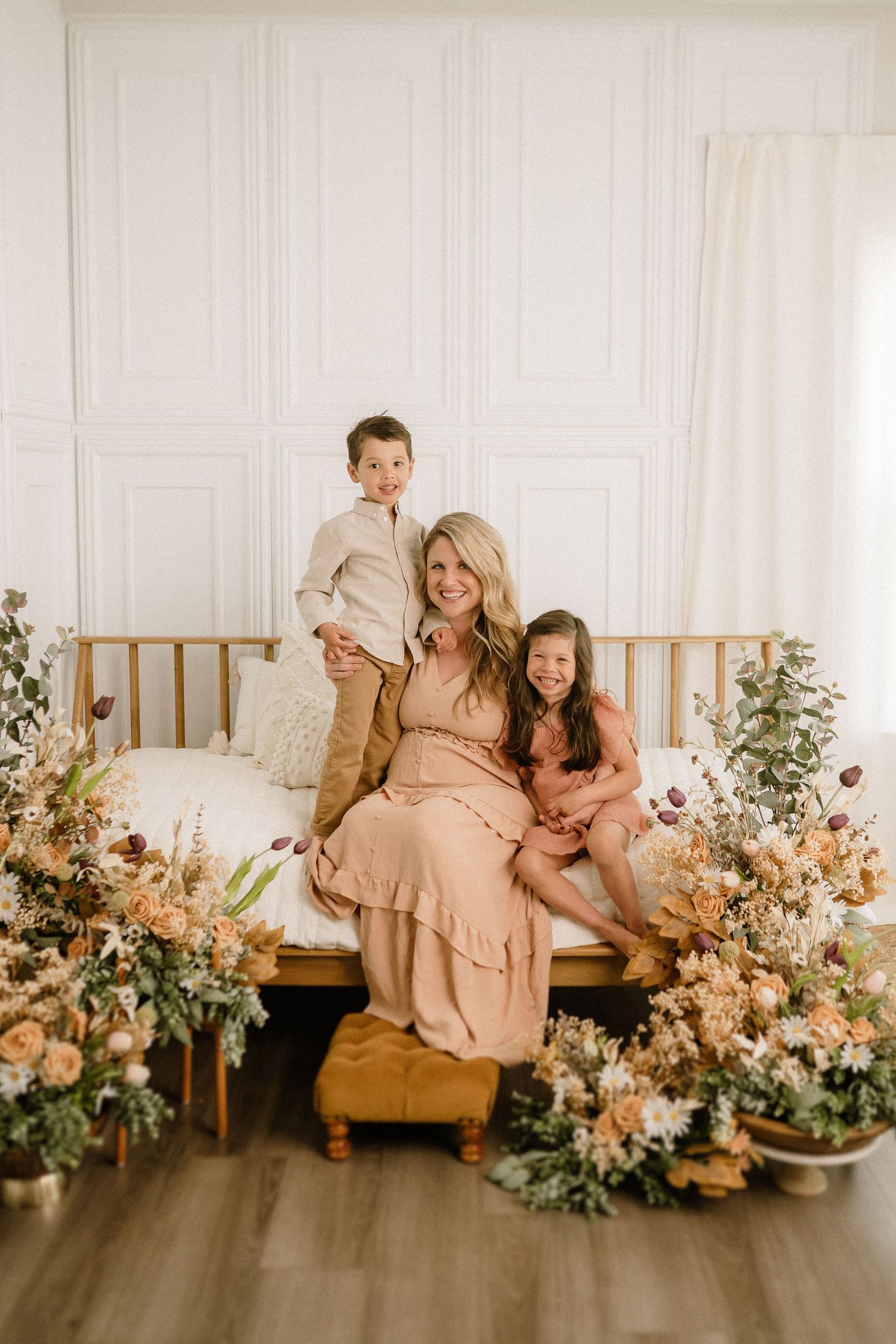 A family sitting on a wooden daybed surrounded by beautiful floral arrangements in a bright white room.