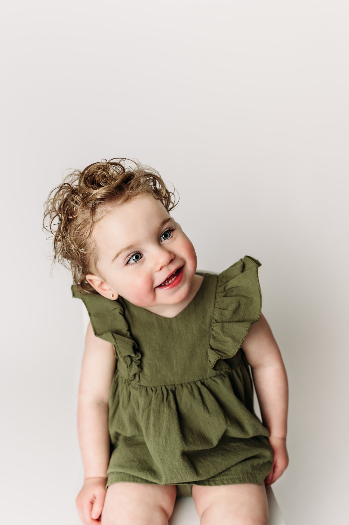 Child in ruffled green dress smiles while sitting against light background.