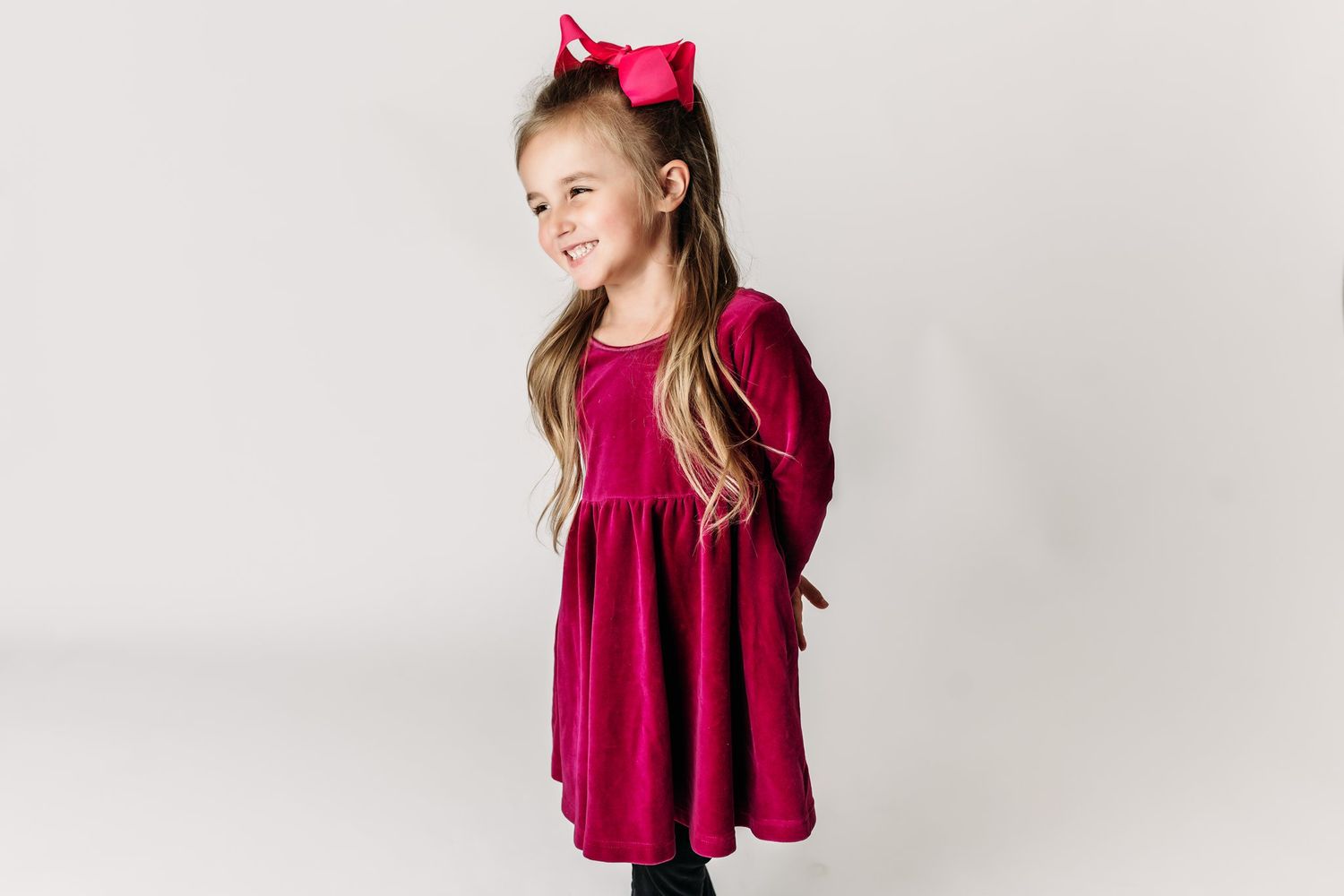 Young model in burgundy velvet dress and pink hair bow stands elegantly against light grey background.