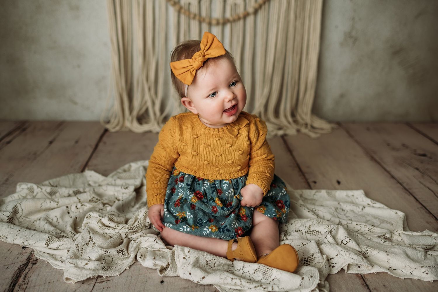 Baby in mustard sweater and floral skirt sits on cream fabric against wooden floor.