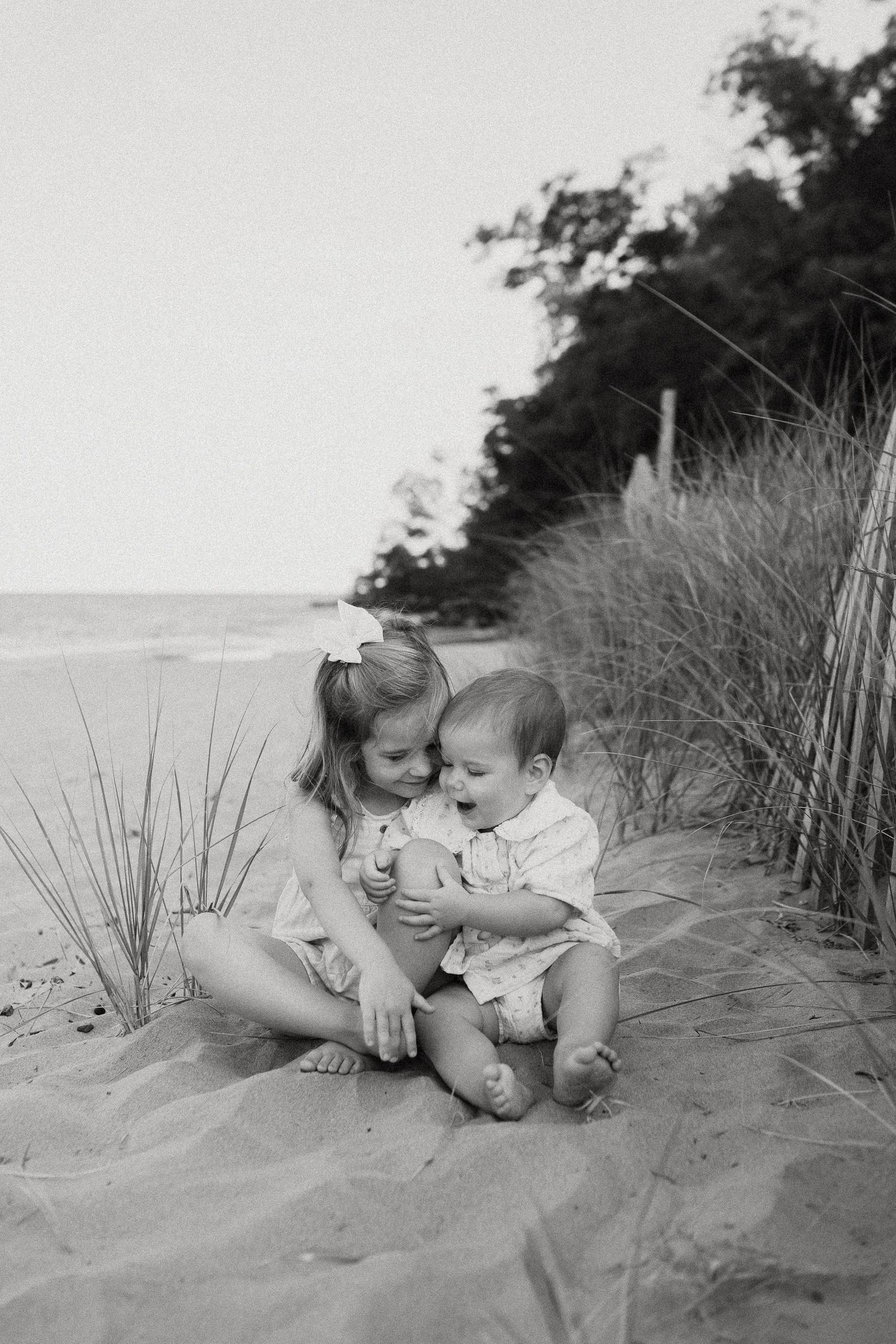Black and white photo of siblings sitting on sandy beach dunes.