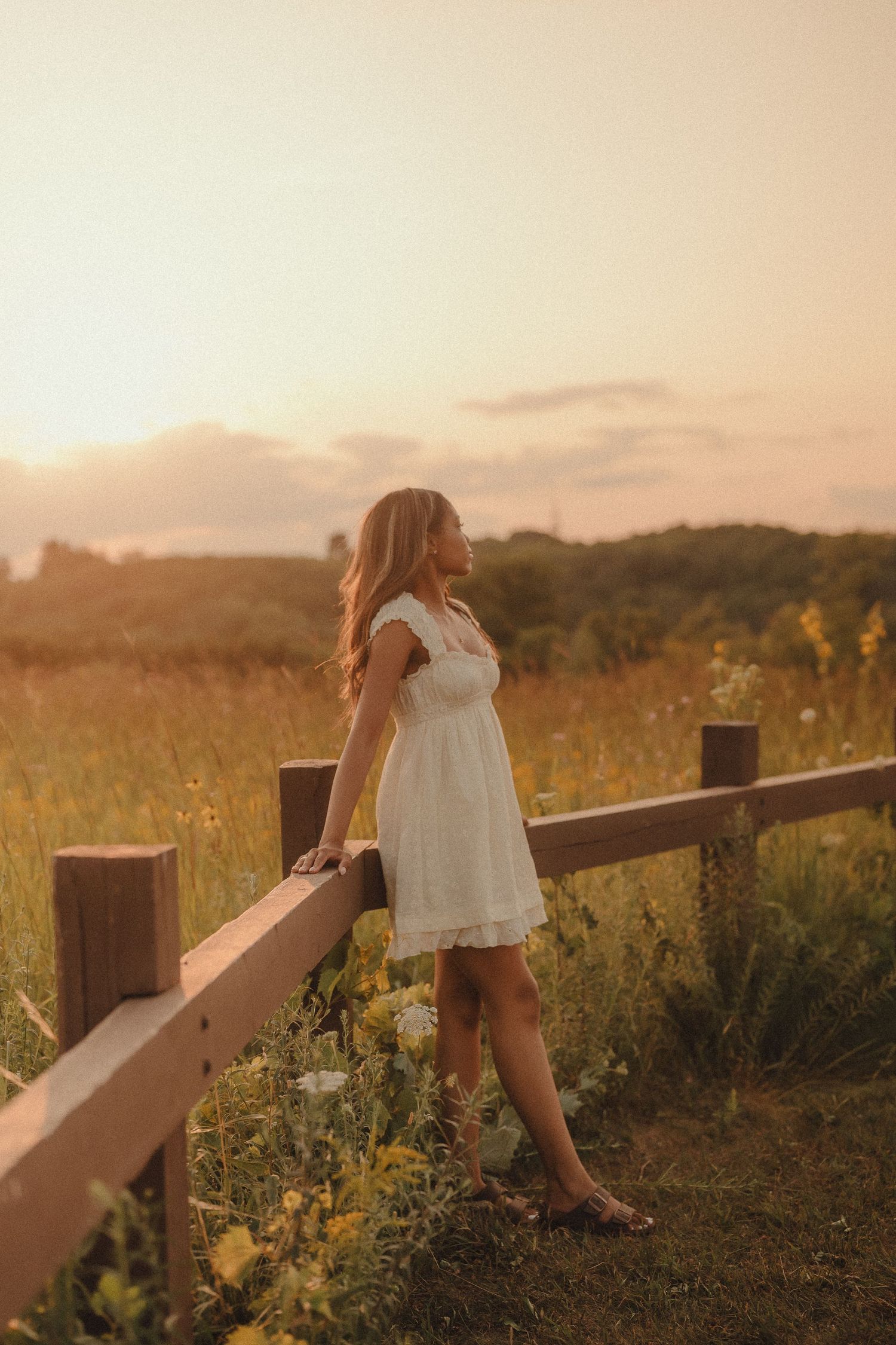 Young woman in white dress standing by rustic wooden fence gazing into a golden sunset field.