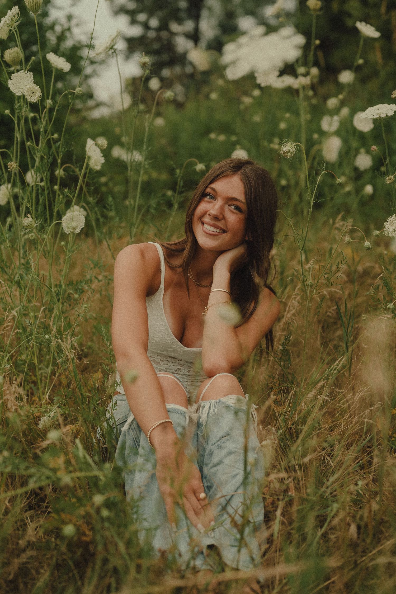 Smiling young woman in white tank top sitting in a wildflower meadow surrounded by tall white flowers.