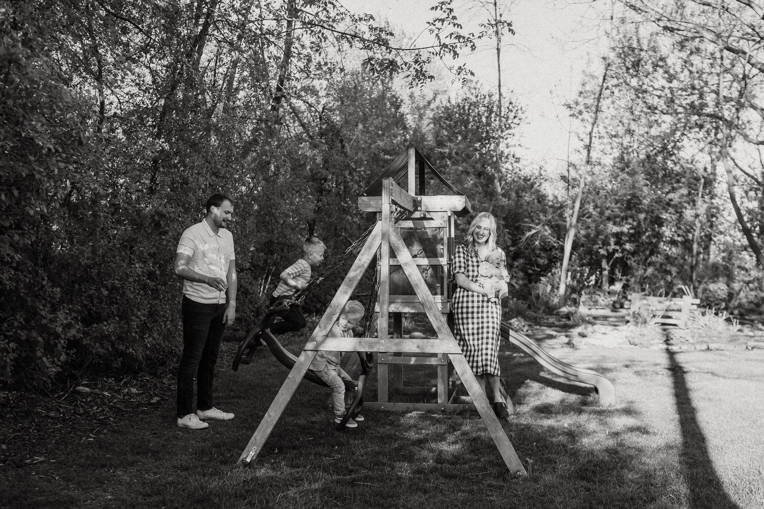 Black and white photo of a family assembling a wooden swing set in a backyard surrounded by trees.