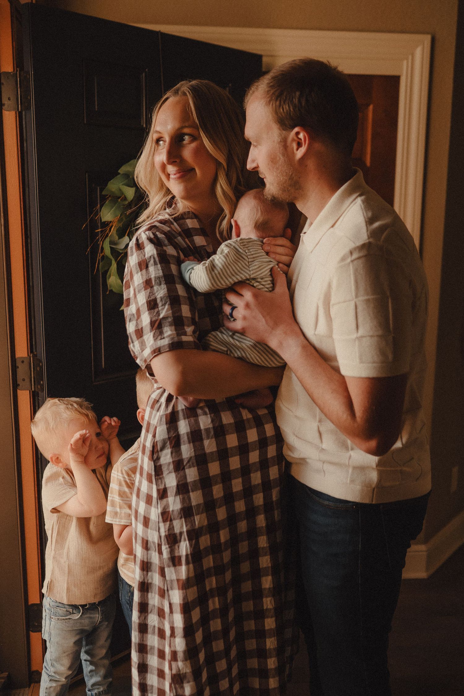 Parents in home entryway admiring newborn baby while young toddler stands beside them smiling.