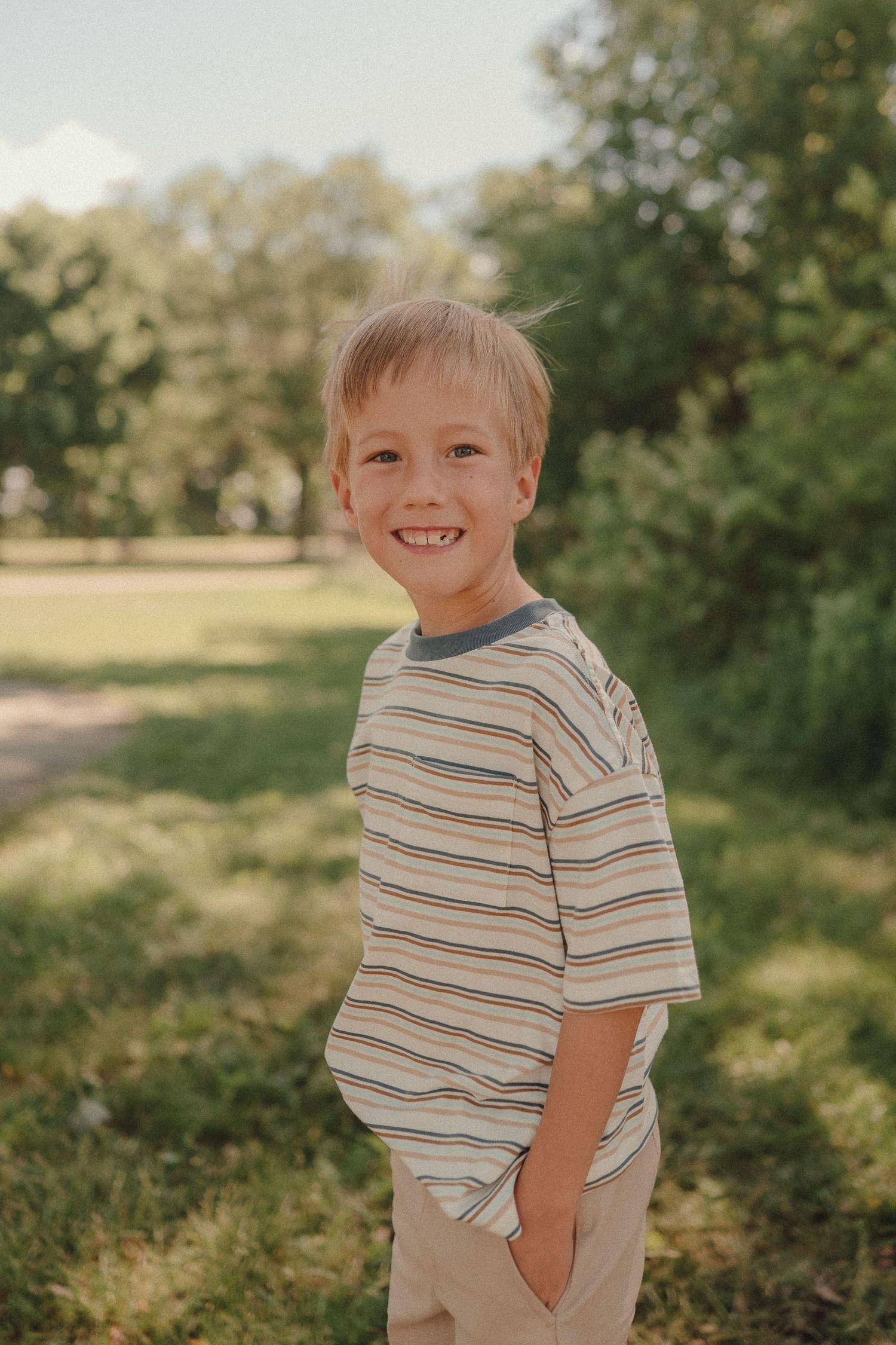 A smiling young blonde boy in a striped t-shirt stands with hands in pockets in a green park.