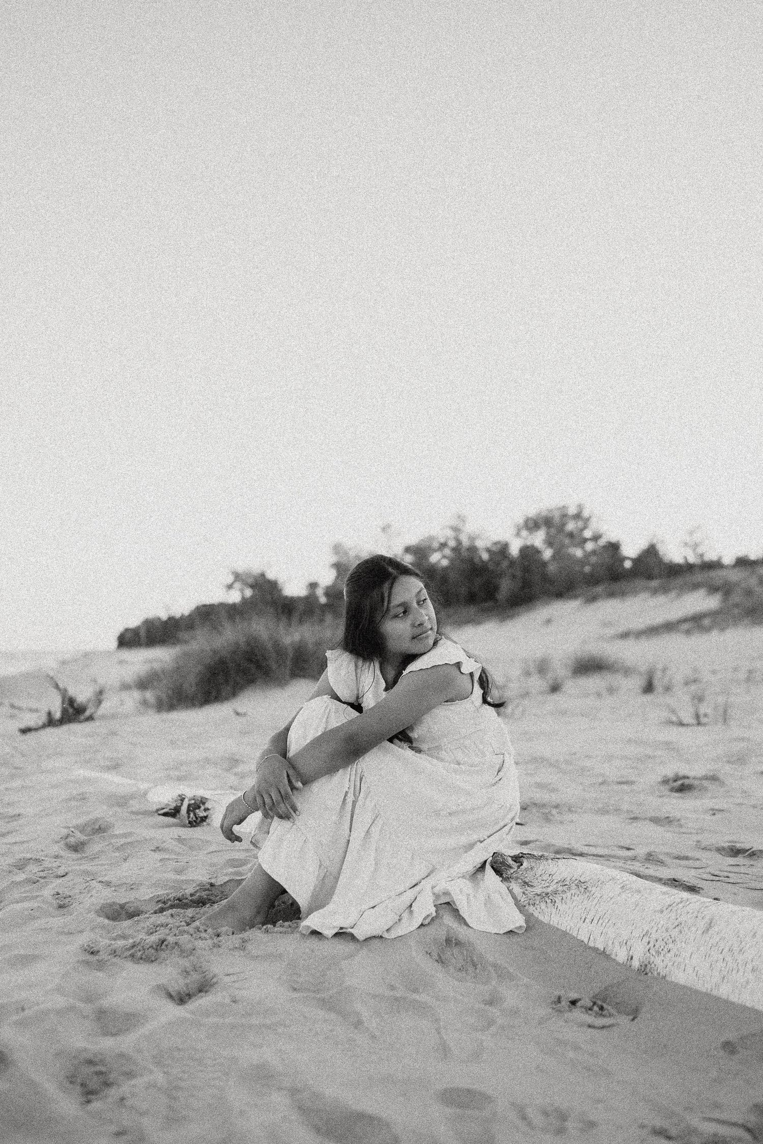 Young woman in white dress sitting on sand dunes in serene black and white portrait photography.