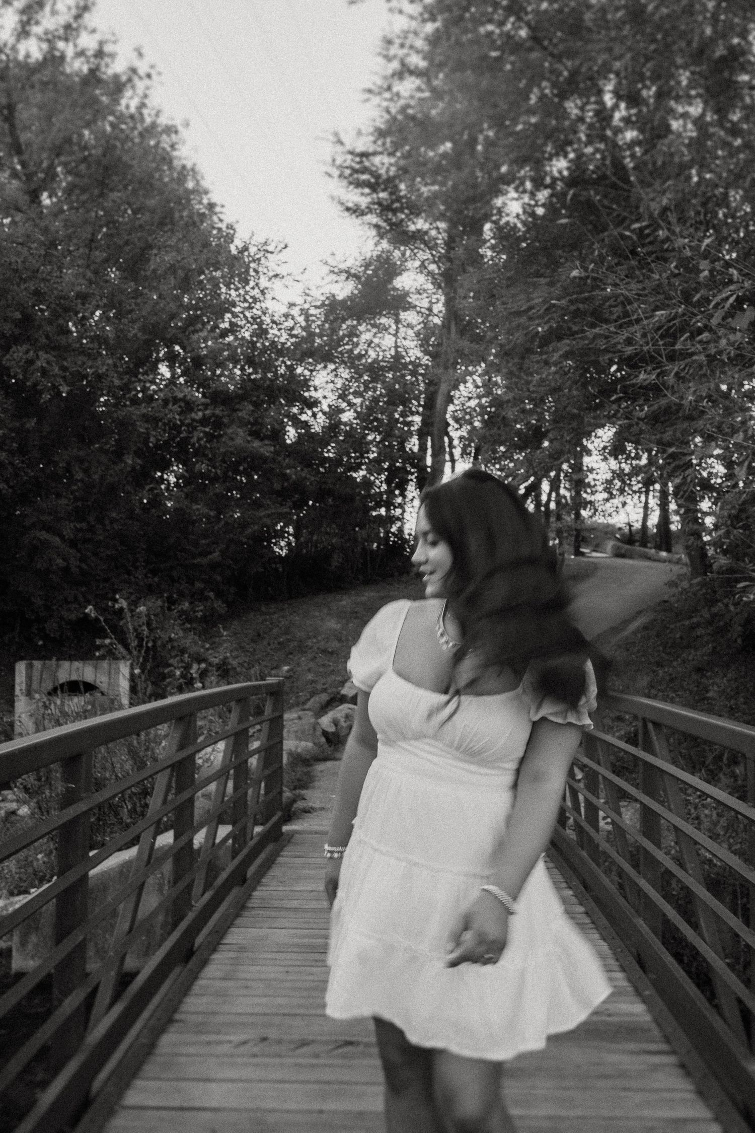 A candid black and white shot of a person in a white dress on a wooden bridge surrounded by trees.