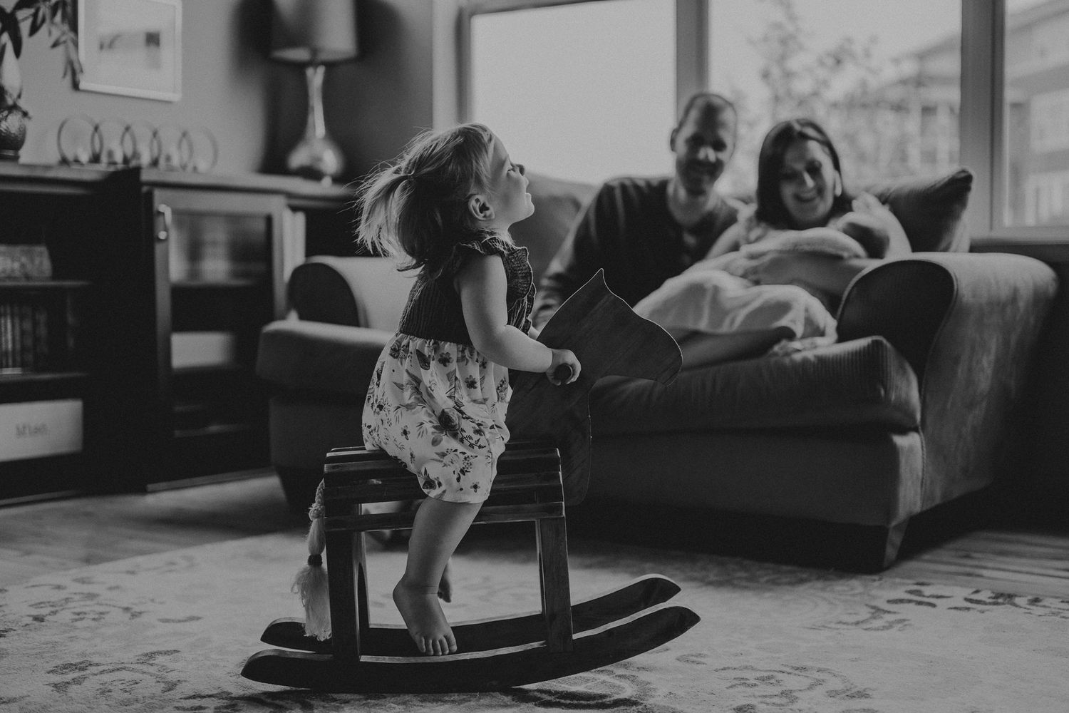 A small child plays on a wooden rocking horse while others relax on a sofa in the background.