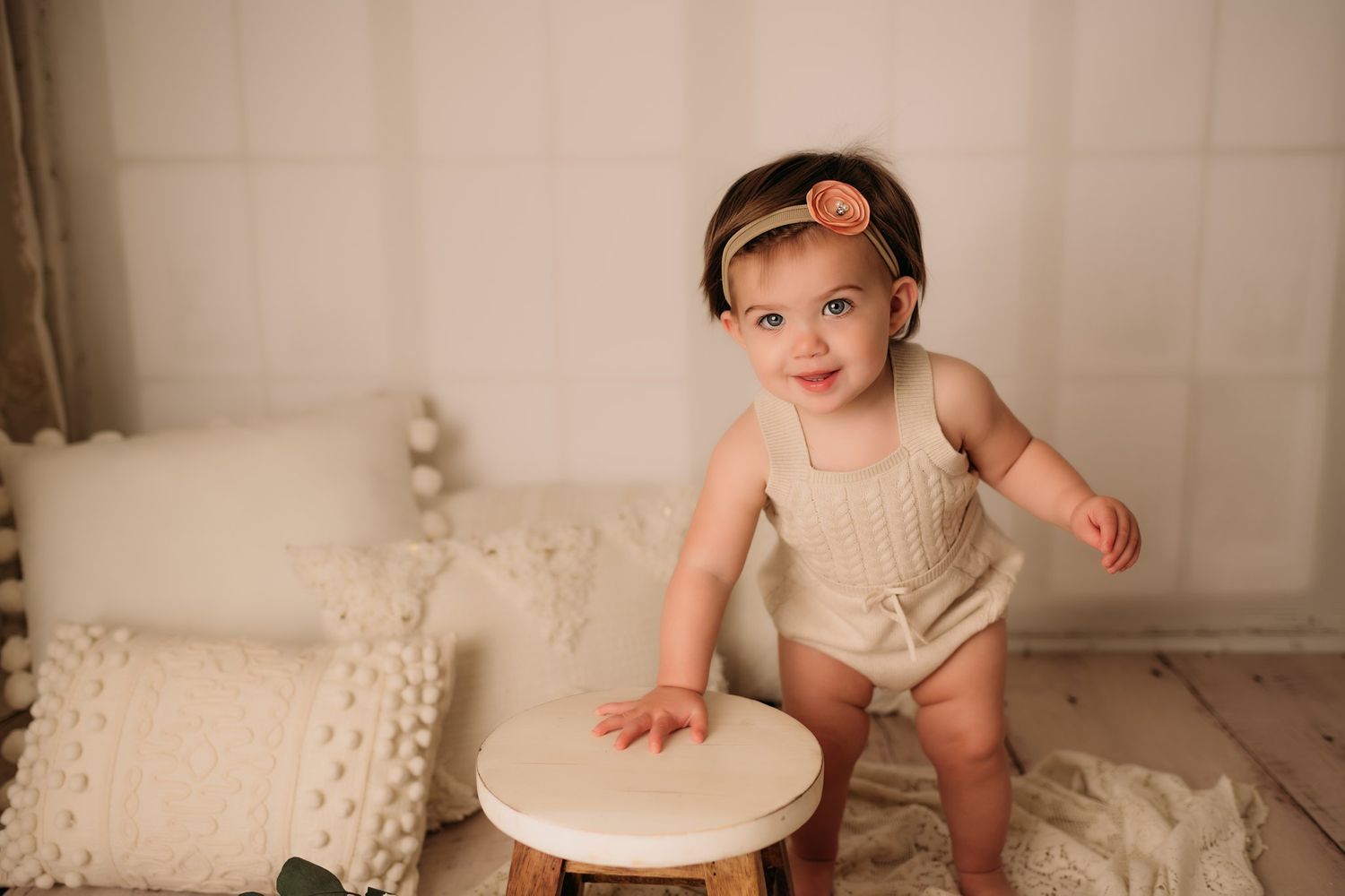 Small baby in beige romper and headband sitting at white wooden table on cream textured blankets in studio setting.