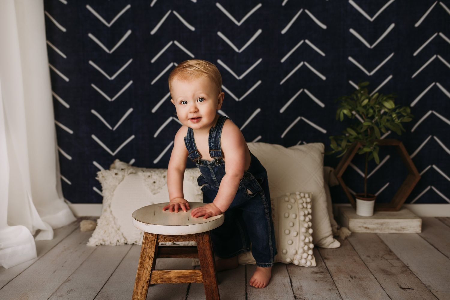 Baby in denim overalls stands near wooden stool against navy geometric wallpaper.
