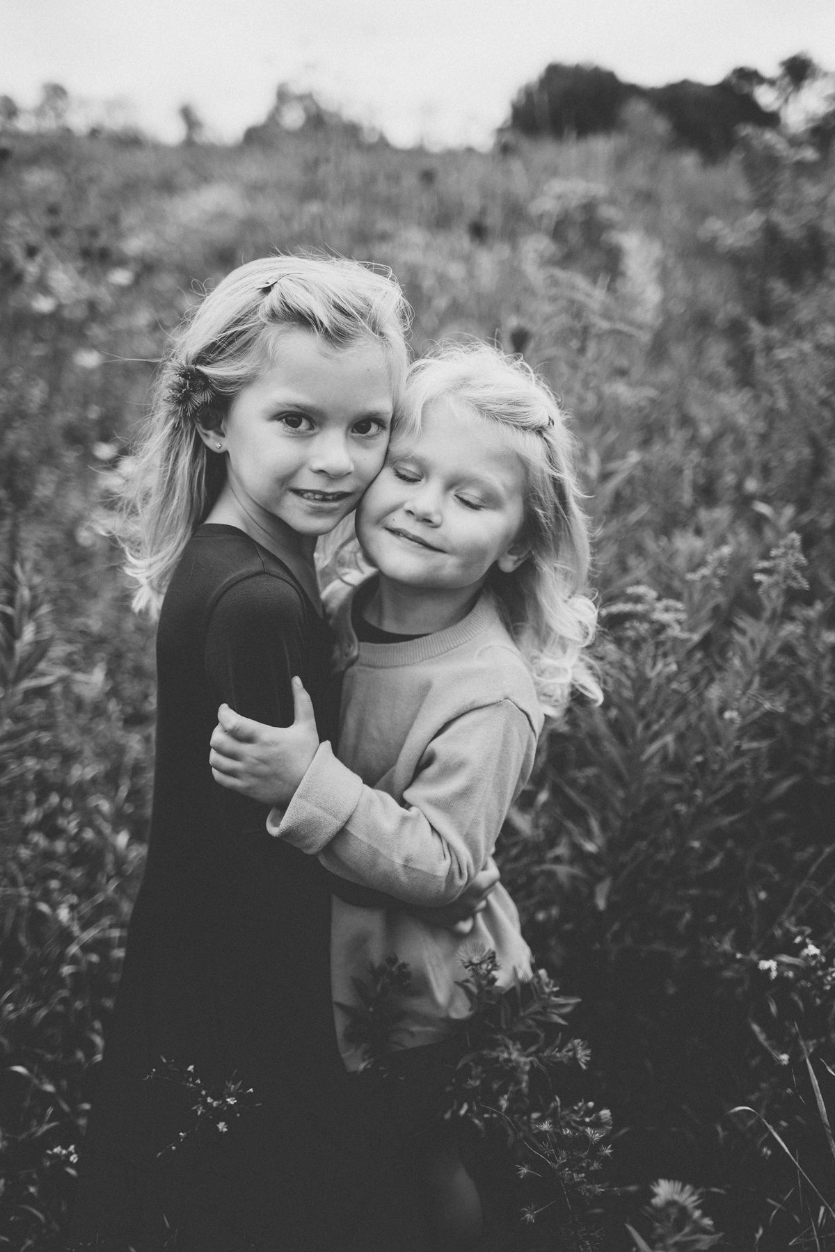 A black and white portrait of two children embracing in a meadow.