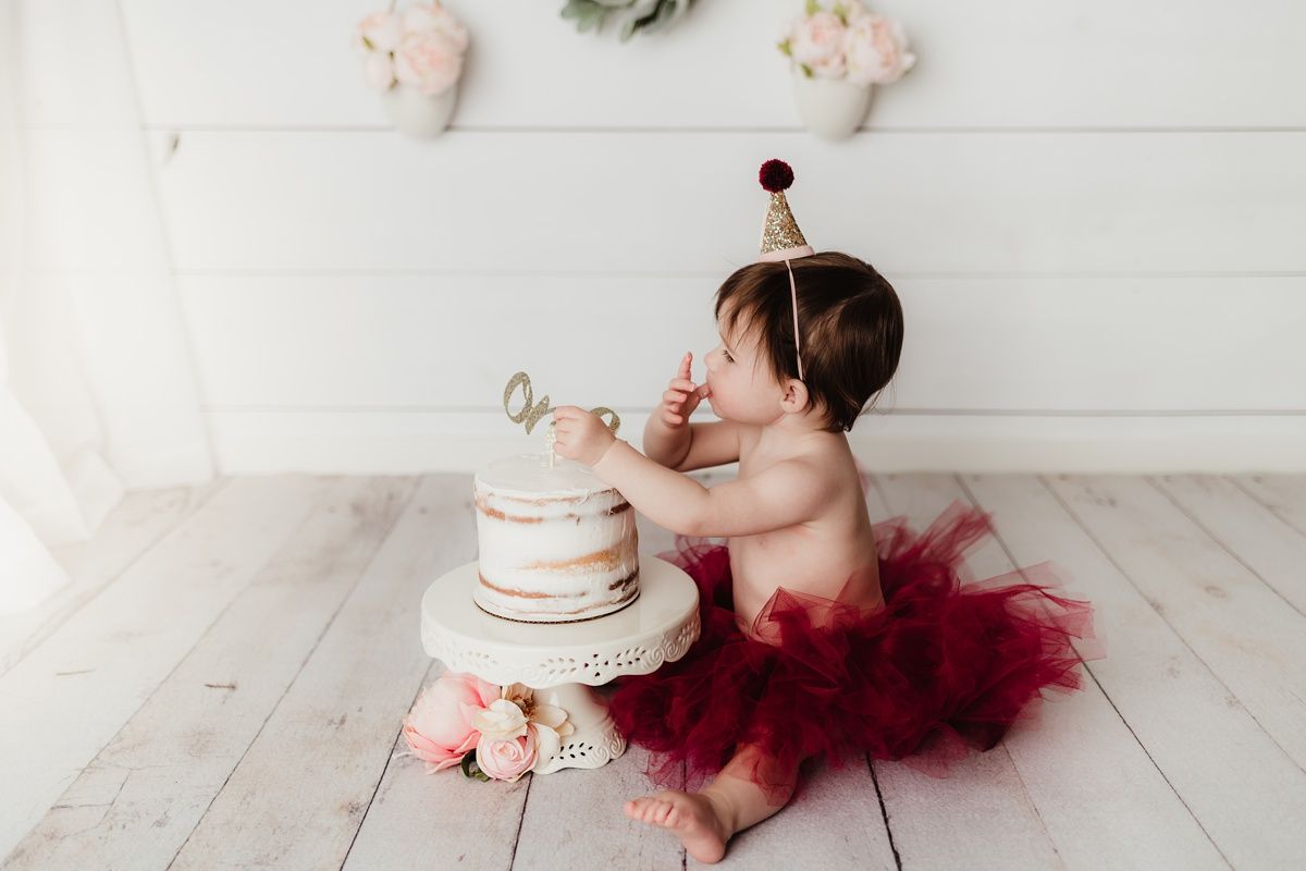 Baby in red tulle skirt reaches excitedly for birthday cake during photo session.