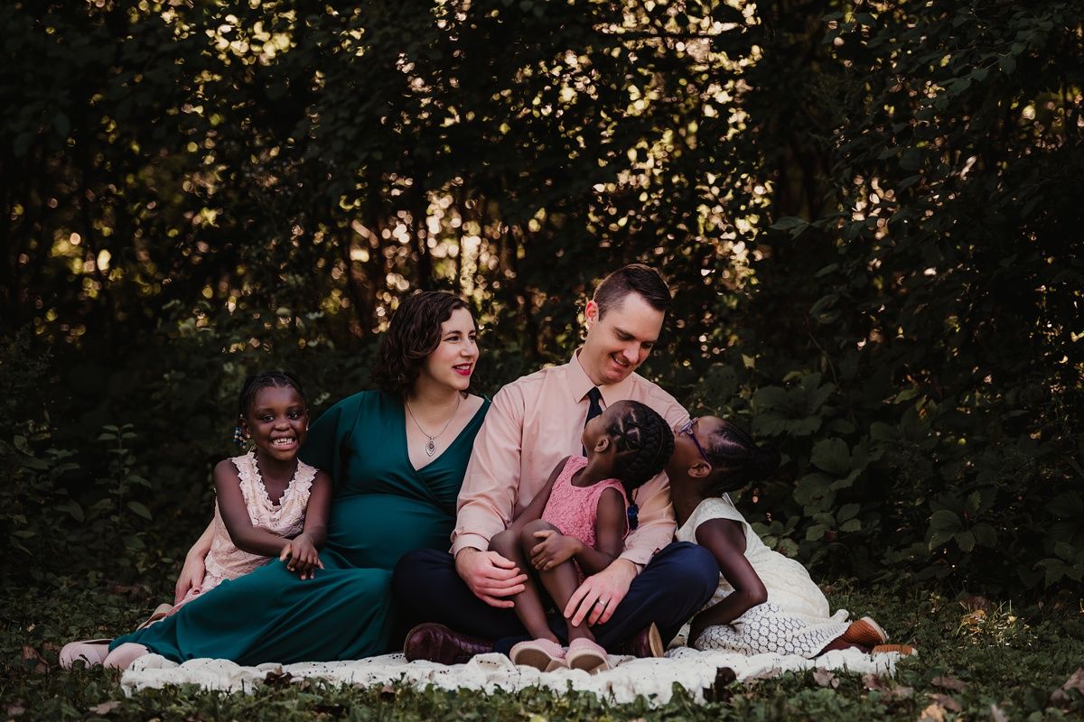 Family relaxes together on blanket during outdoor photo session in wooded setting.