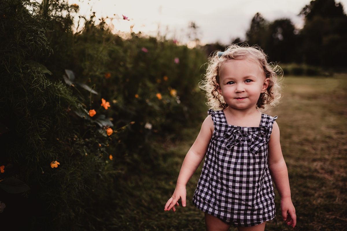 Happy toddler in checkered dress playing outdoors near flowering bushes.