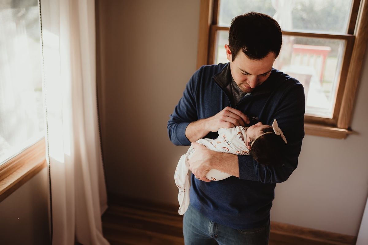 Parent in navy sweater tenderly holds sleeping newborn by window.