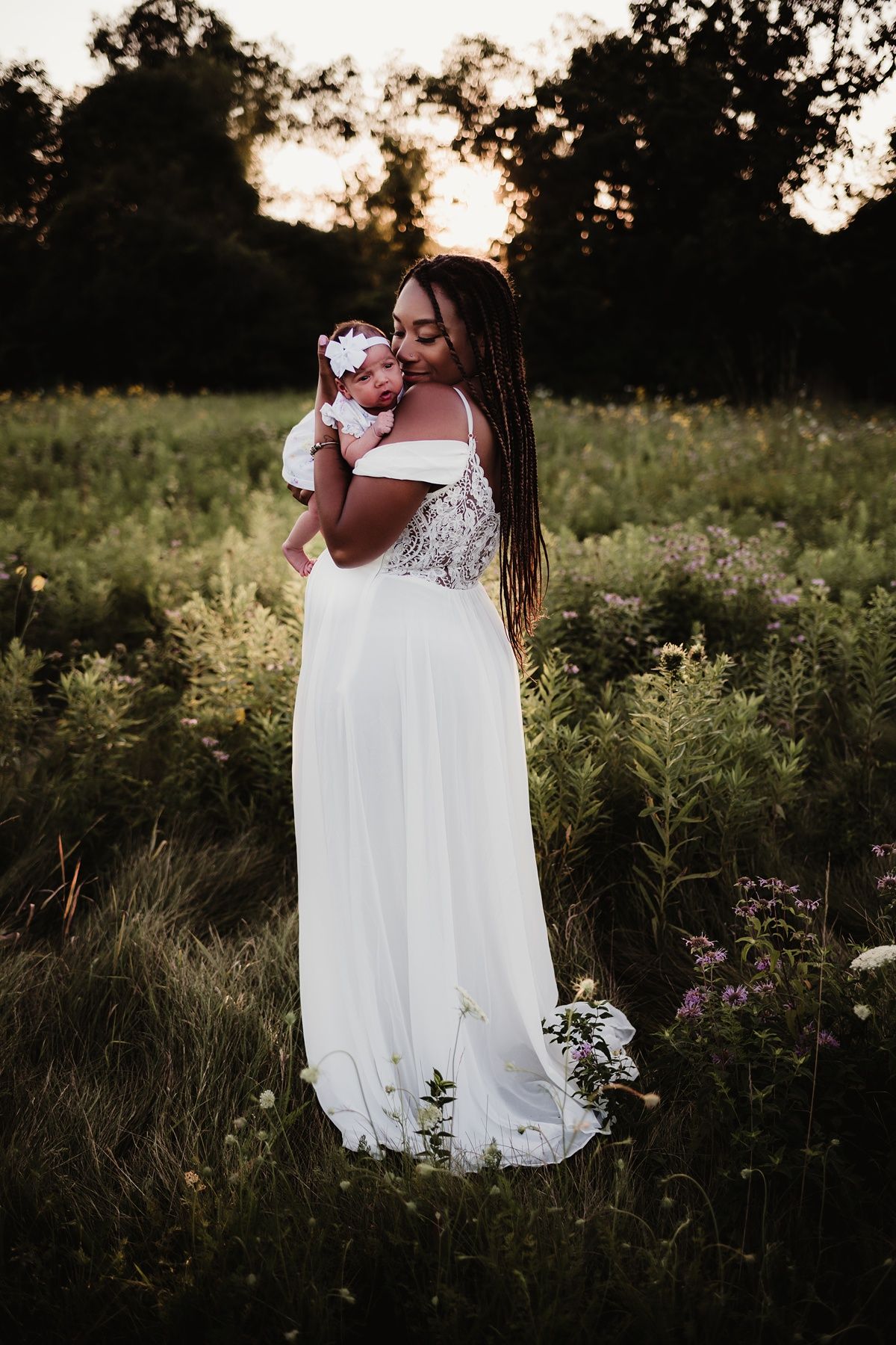 A woman in a lace white dress cradles a baby among wildflowers at sunset.