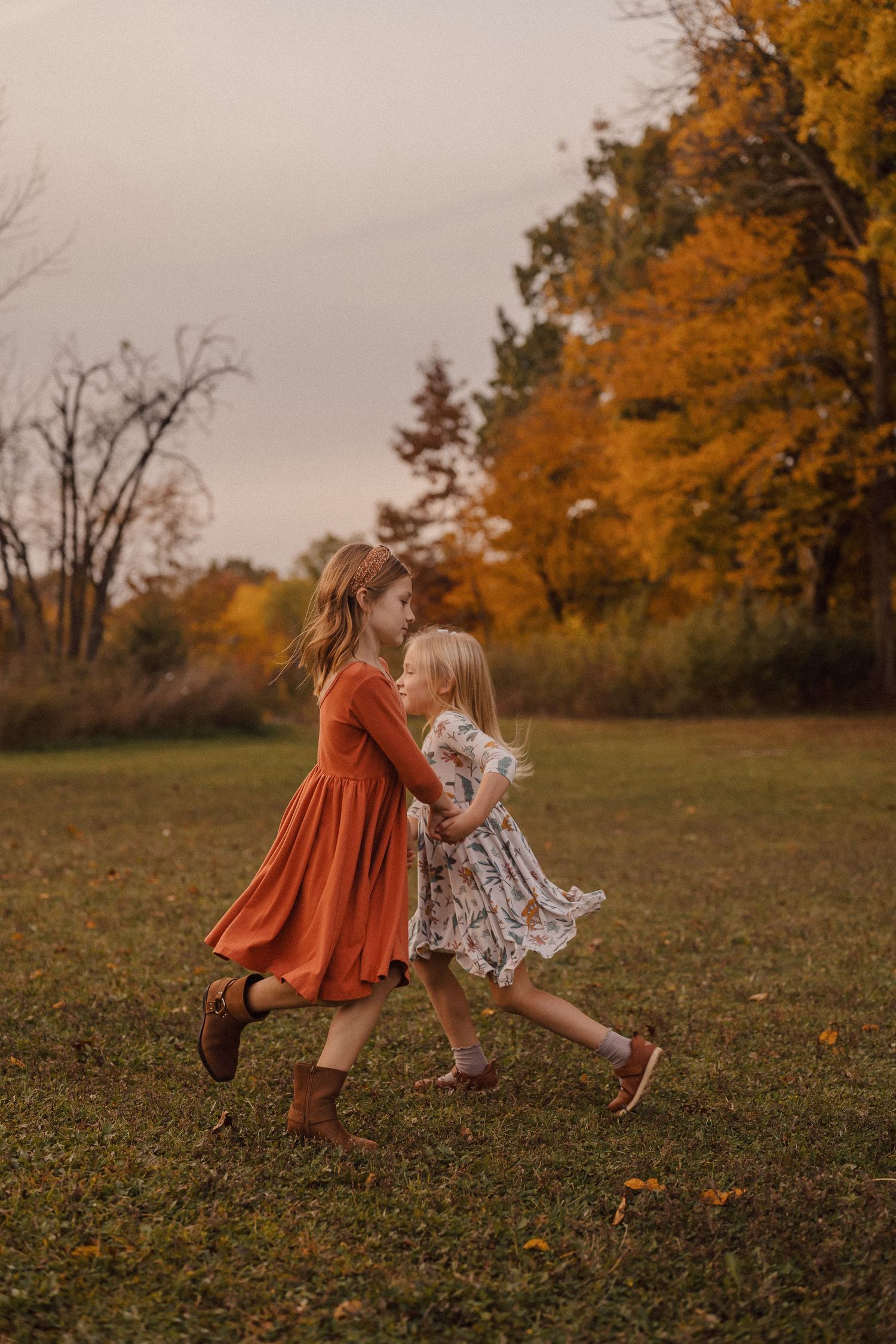 Children in autumn dresses dance and twirl in a field with golden fall trees in the background.