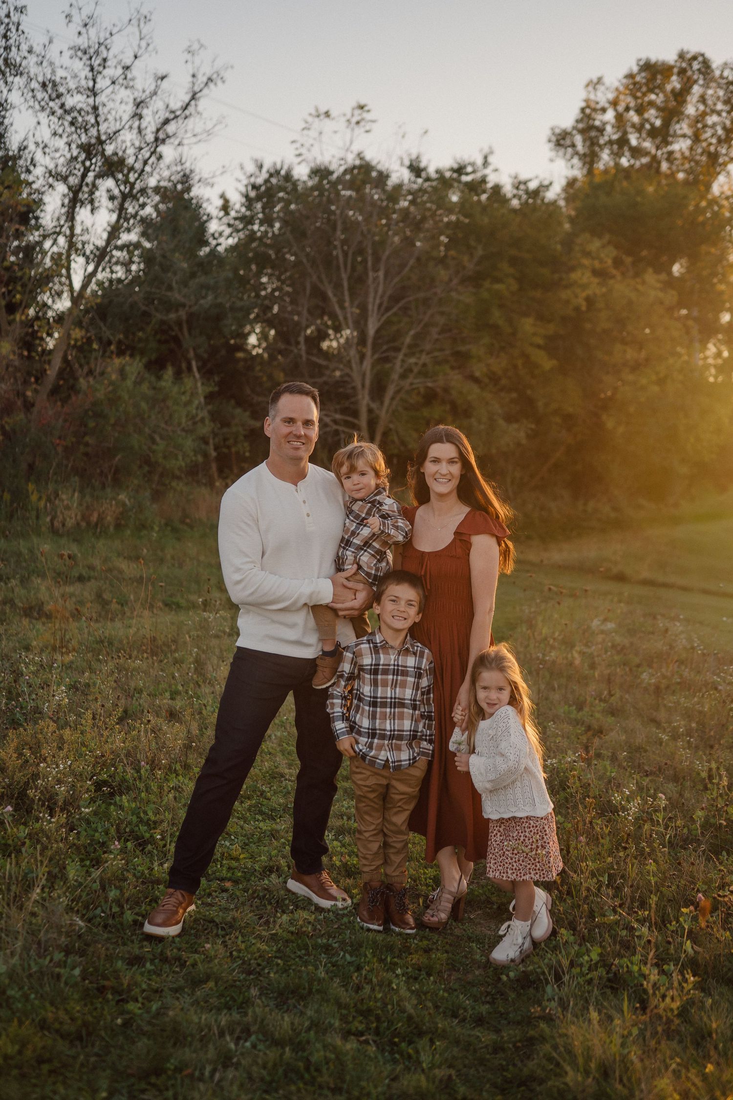A family portrait captured in a rustic outdoor setting during golden hour with trees in the background.