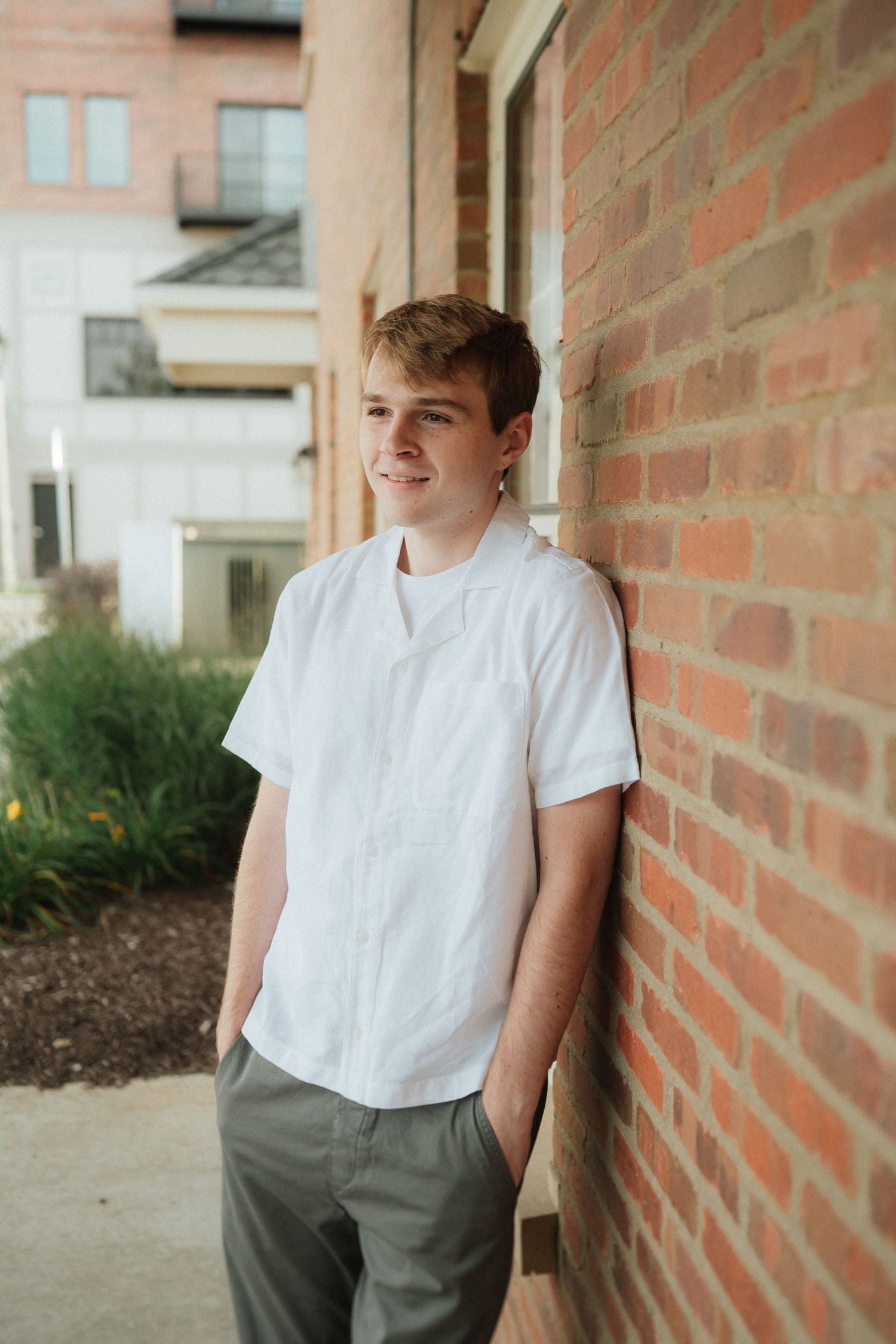 Person in white t-shirt and gray pants poses casually against brick wall in natural lighting.
