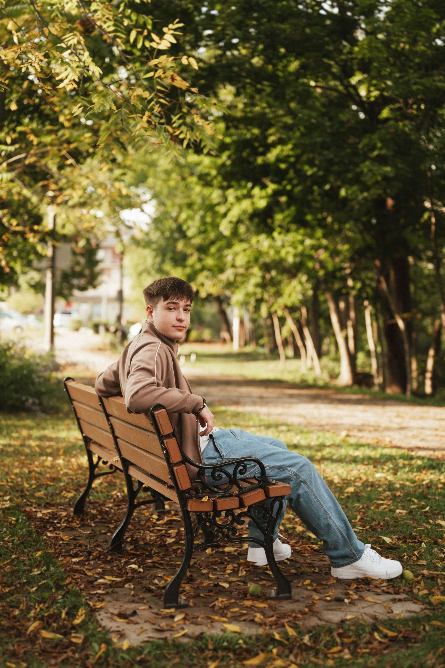 A person sits alone on a wooden park bench surrounded by autumn leaves and trees in golden sunlight.