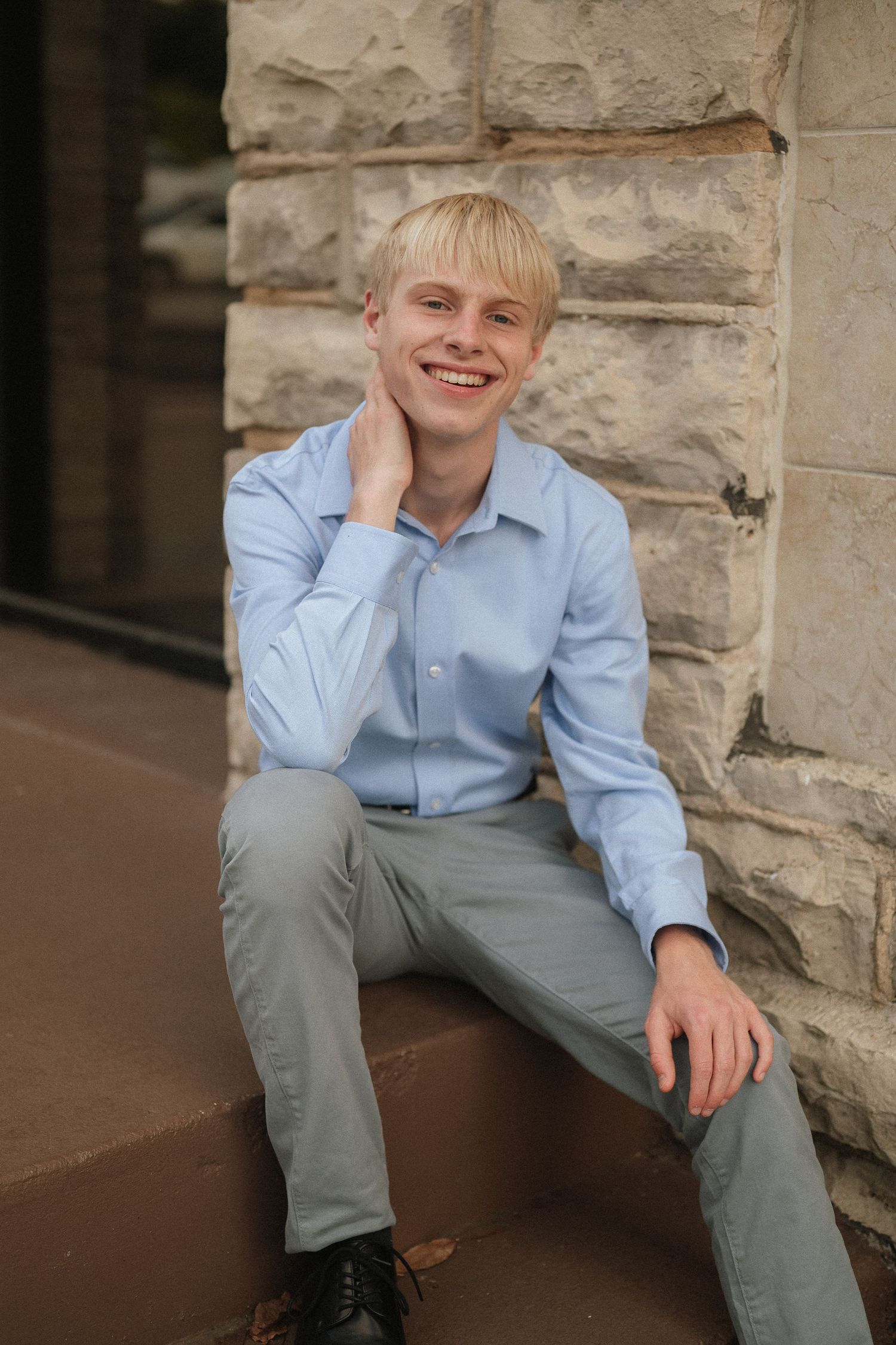 A casual portrait shows someone seated on a stone ledge wearing a light blue shirt and gray pants.