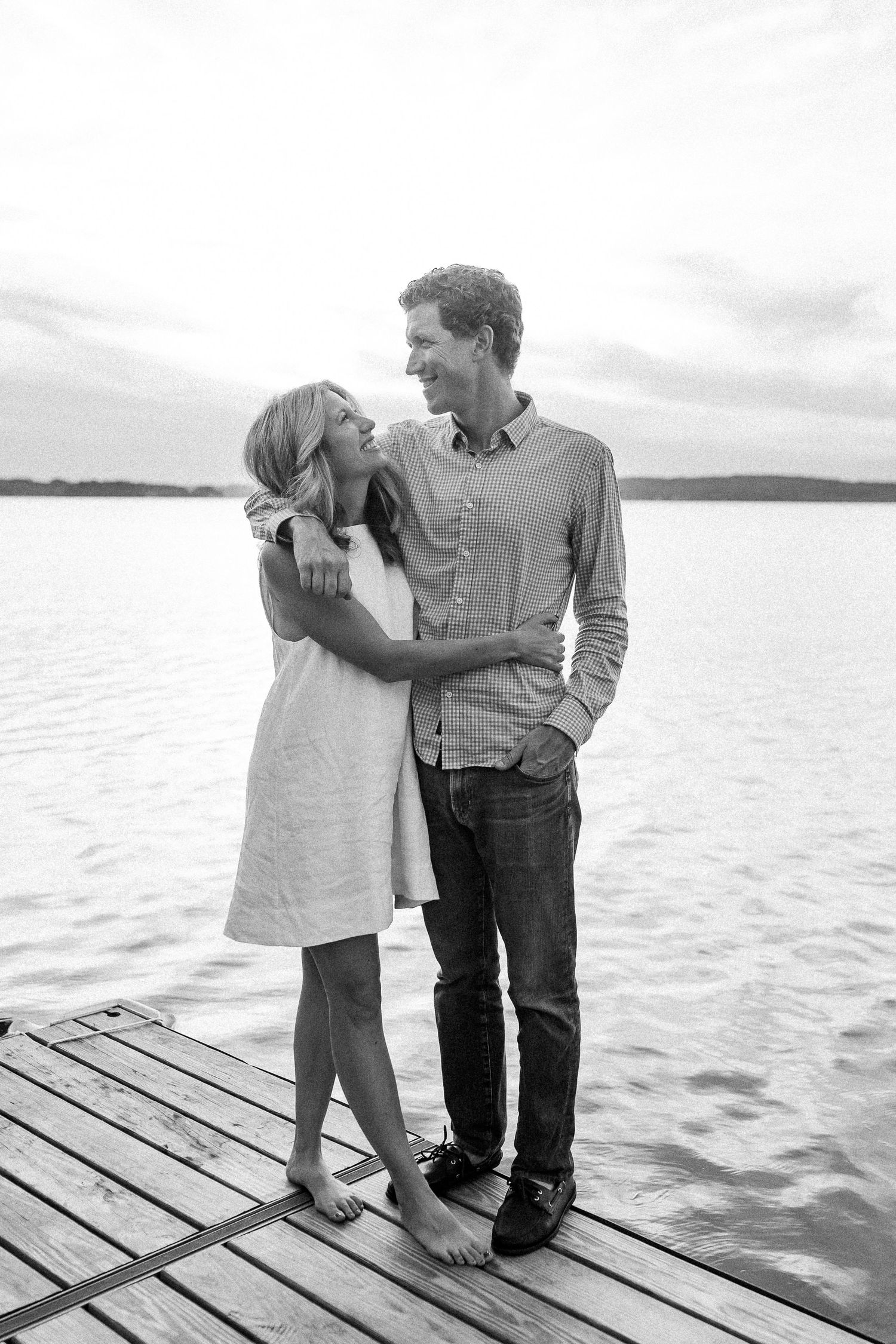A couple stands together on a wooden dock overlooking a serene lake on a cloudy day.