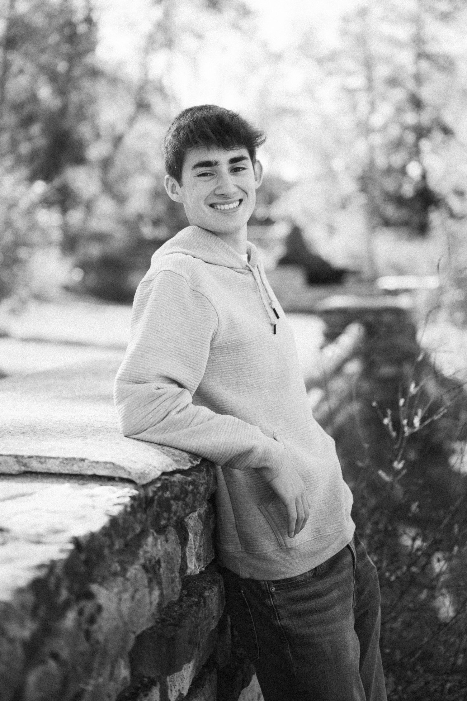 Black and white portrait of someone sitting on a stone wall with trees in background.