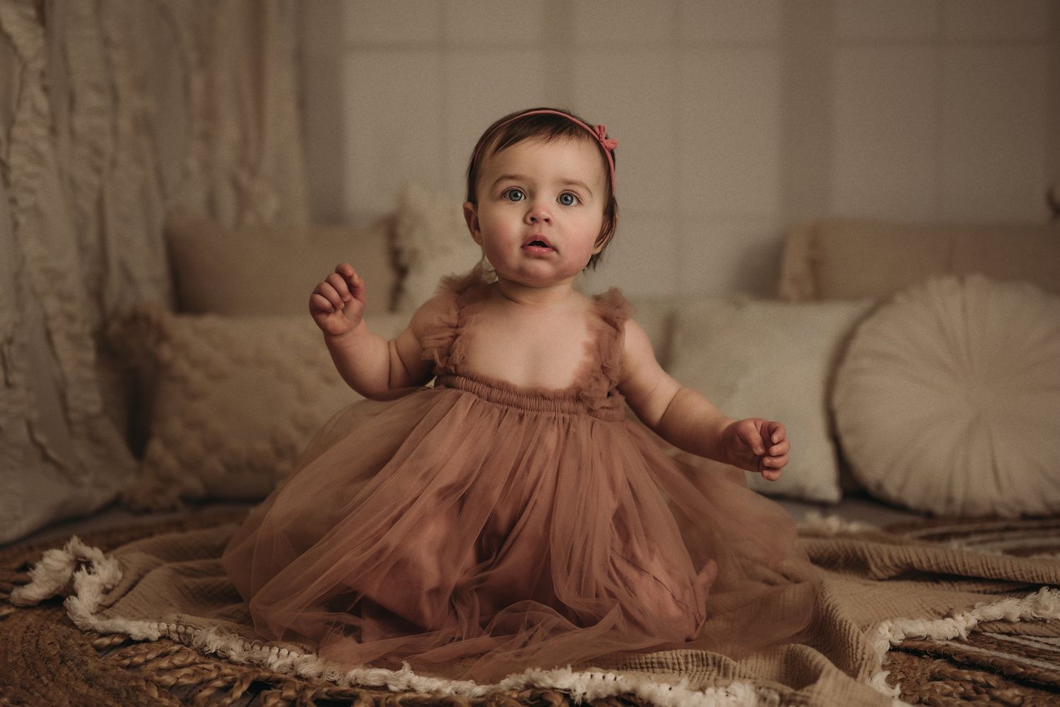 Baby wearing dusty rose tulle dress sitting on textured rug in warm-toned nursery setting.