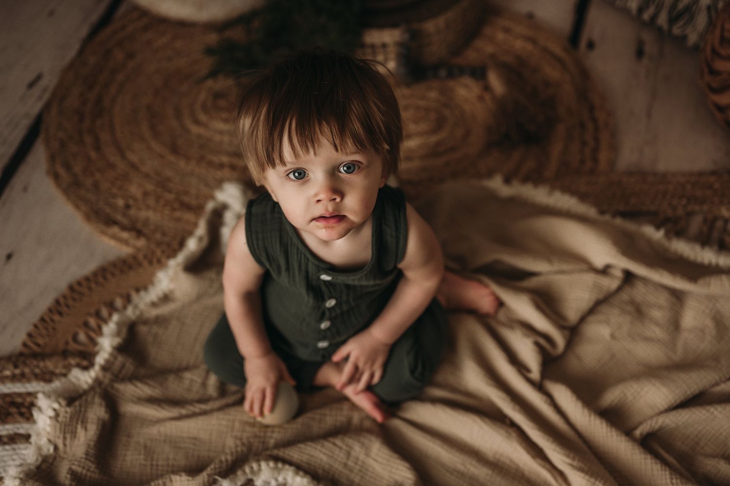 Child in green overalls sits on woven rug with macrame wall hanging in background.