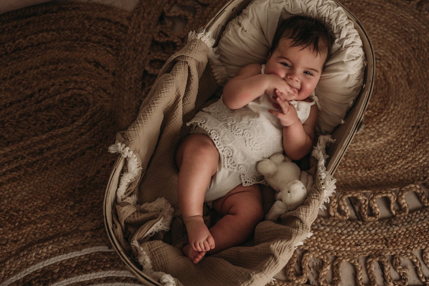 Baby lying in woven bassinet holding plush toy on textured brown rug.