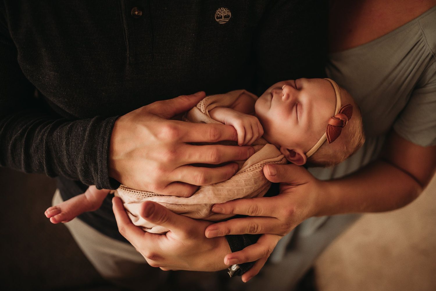 A sleeping newborn swaddled in beige fabric is tenderly held in gentle hands.