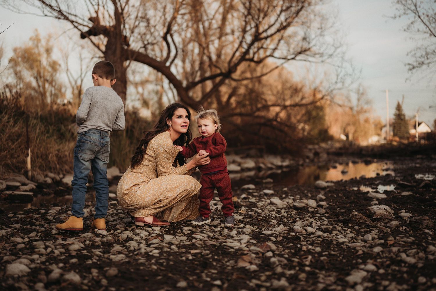 Candid family moments captured by stream with autumn trees and golden light in background.
